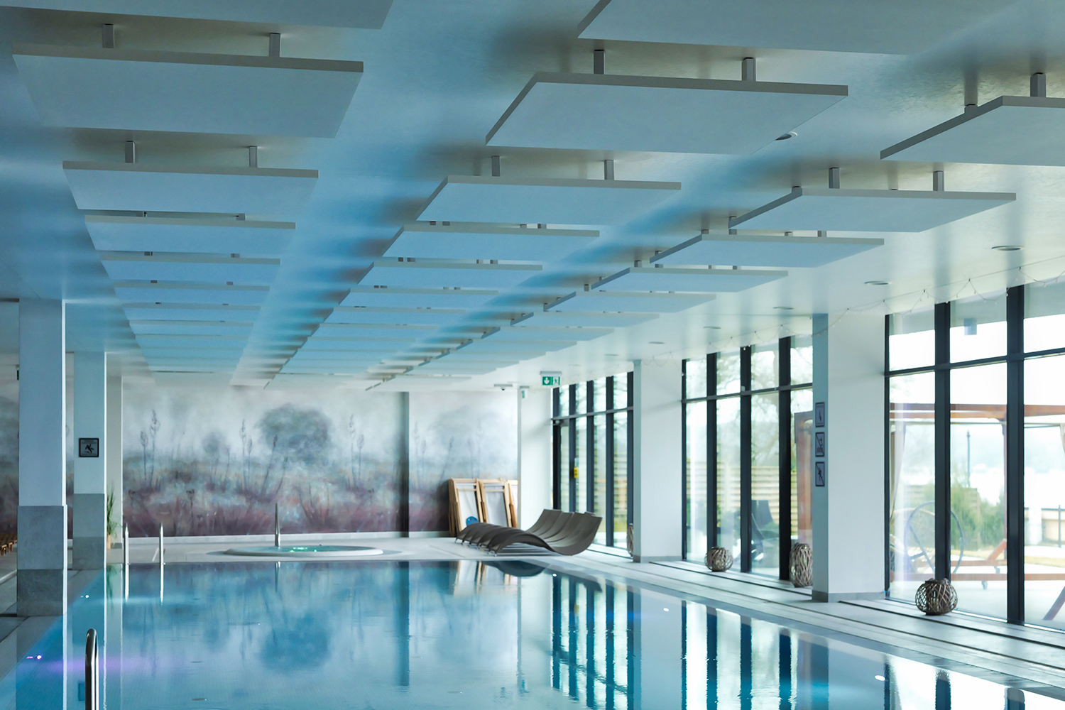 Indoor swimming pool area with large windows letting in natural light, modern ceiling panels, loungers, and a hot tub at the far end. The space has a clean, contemporary design.