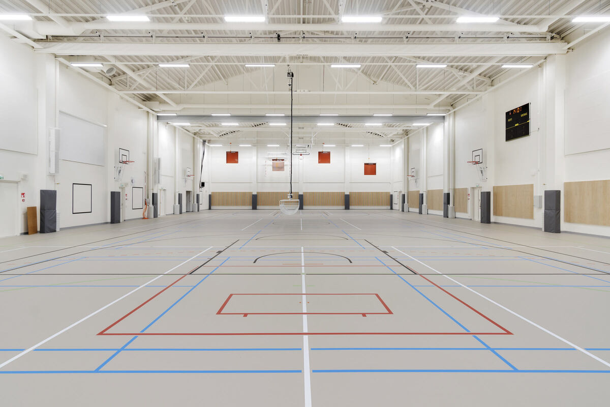 Wnętrze dużej, pustej sali gimnastycznej z kilkoma boiskami do koszykówki oznaczonymi czerwonymi i niebieskimi liniami. Sufit jest wysoki z widocznymi belkami, a na ścianach zamontowane są kosze i tablica wyników po prawej stronie.