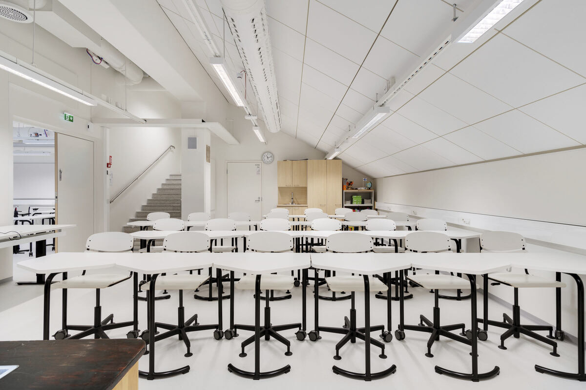 Pusta sala lekcyjna z białymi krzesłami i biurkami ustawionymi w rzędach. Pomieszczenie ma skośny sufit, świetlówki i zegar ścienny. W tle widać schody prowadzące na wyższy poziom.