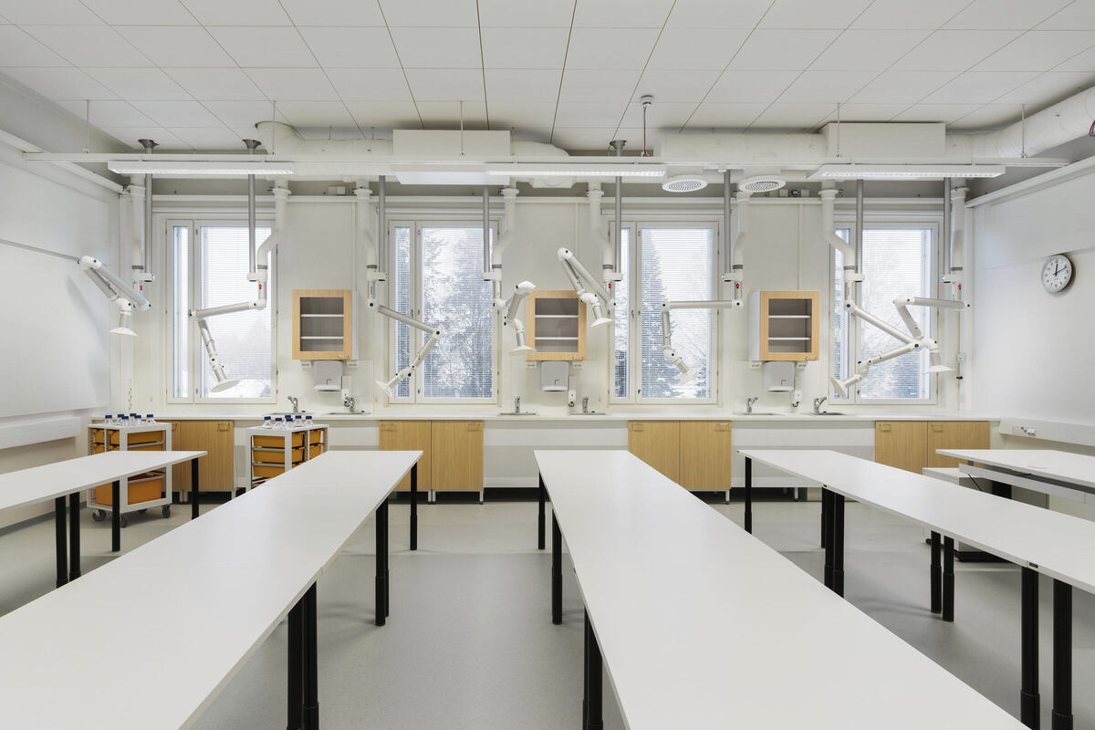 Oto tłumaczenie:  Nowoczesne laboratorium naukowe z długimi białymi stołami, drewnianymi szafkami i sprzętem wentylacyjnym. Kilka okien pozwala na naturalne oświetlenie przestrzeni. Okrągły zegar ścienny jest zamontowany blisko sufitu.