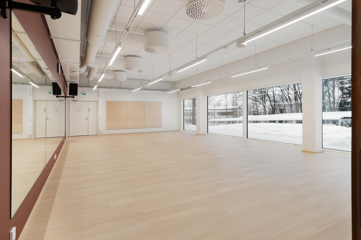 Puste, nowoczesne studio taneczne z jasną drewnianą podłogą, dużymi lustrami na jednej ścianie i oknami po przeciwnej stronie, wpuszczającymi naturalne światło. Na zewnątrz okien widać śnieg. Sufit ma widoczne przewody wentylacyjne i oświetlenie sufitowe.