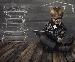 Boy with glasses reading a book in front of a blackboard