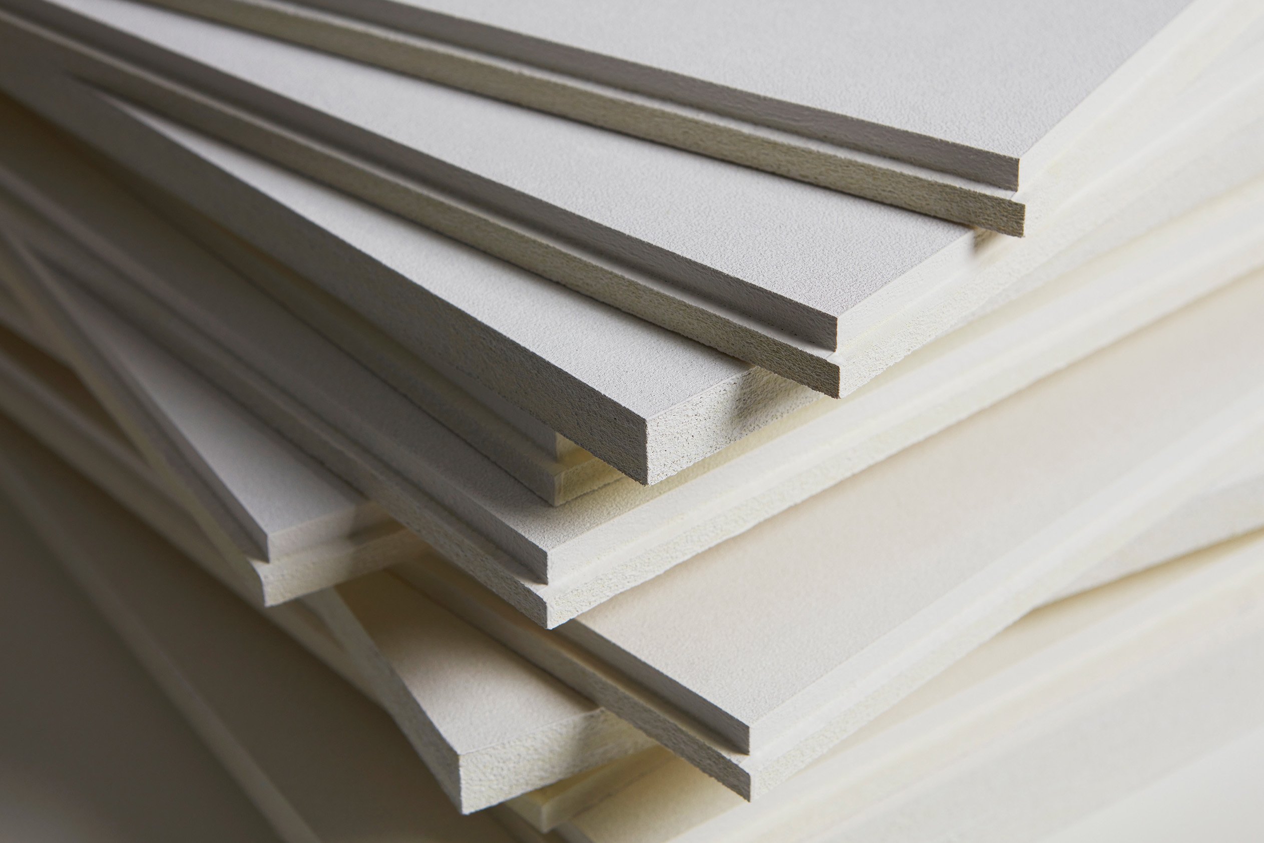A close-up of multiple white acoustic boards stacked on top of each other, showing their thin edges and layered arrangement. The smooth surfaces and precise cuts are visible, creating a geometric pattern.