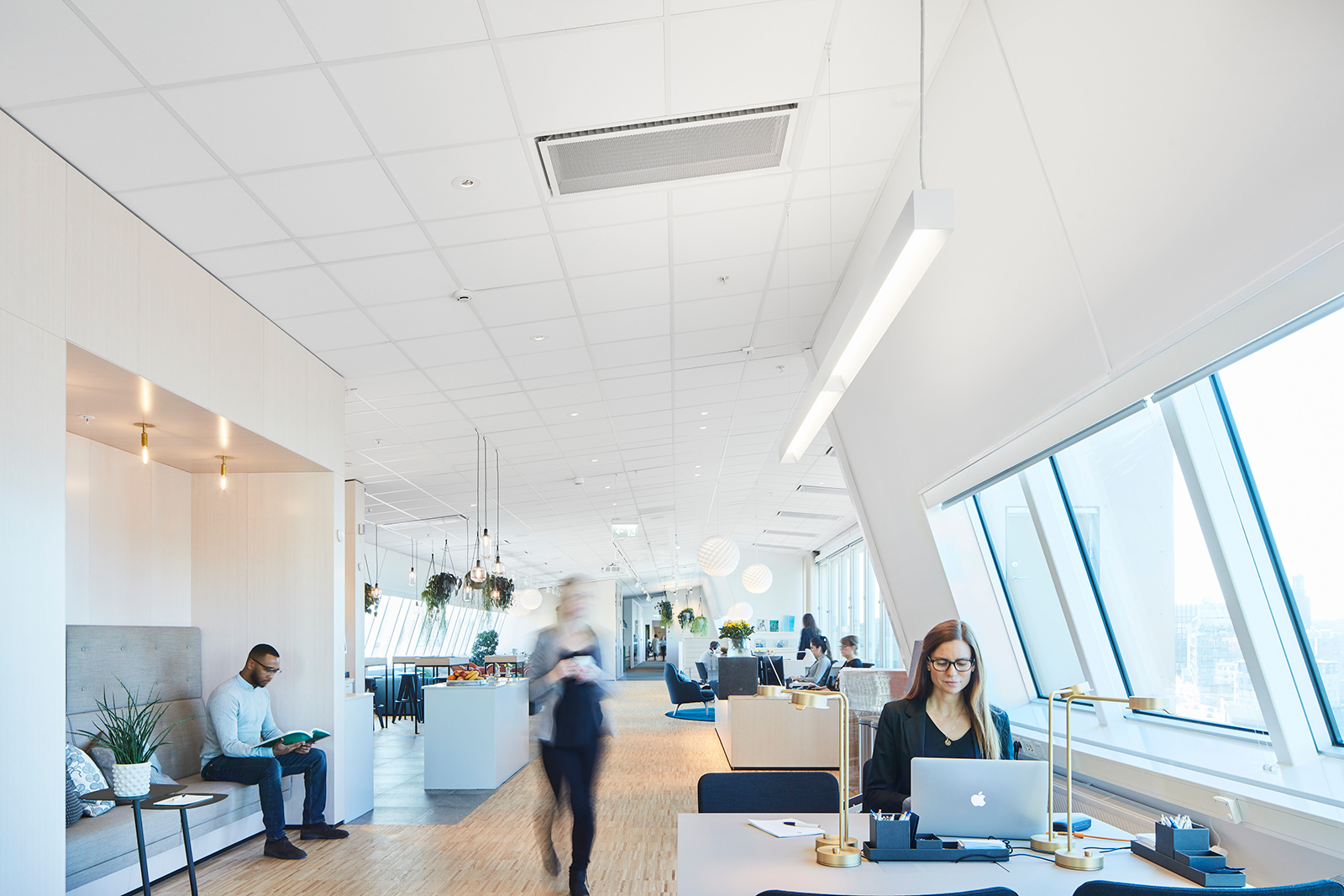 A modern, spacious office with large windows. A woman types on a laptop at a desk in the foreground. Two people sit in a lounge area, while another person walks through the space. The decor is bright and minimalist, with plants and neutral tones.