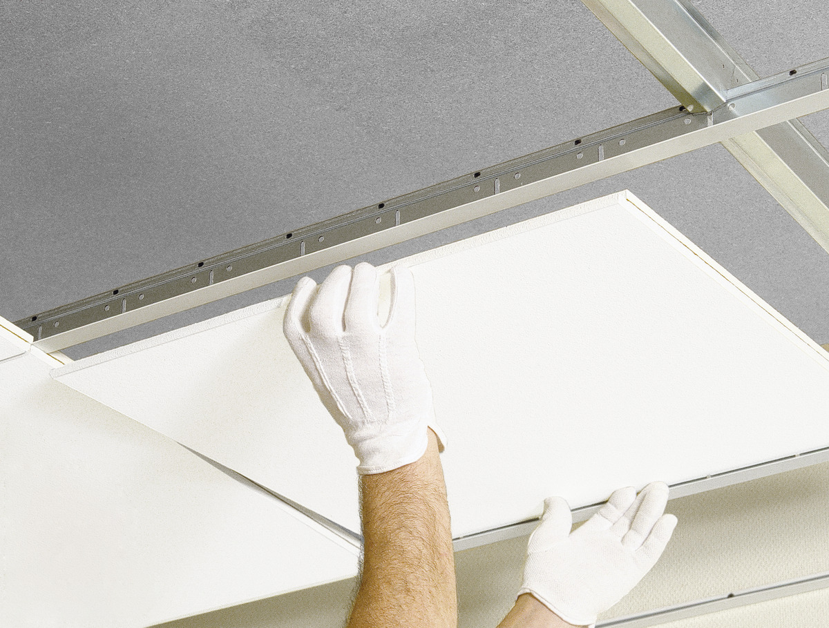 Gloved hands installing a ceiling tile into a metal grid, illustrating a suspended ceiling system. The tile is almost in place against a backdrop of other tiles and the metal framework.