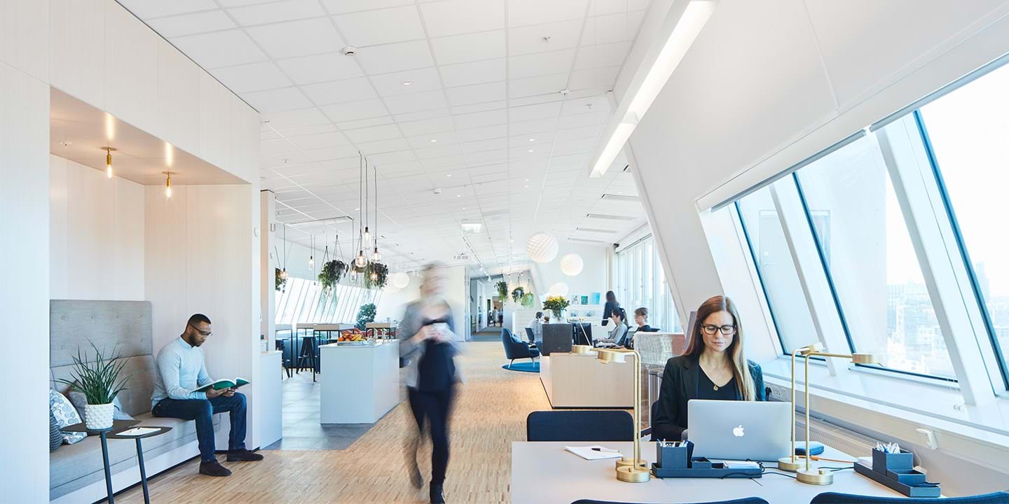 A modern office space with large windows and wooden flooring. A person works on a laptop at a desk, another sits reading a book. Blurred figure walks by. Plants and decorative lights add ambiance to the bright, open environment.