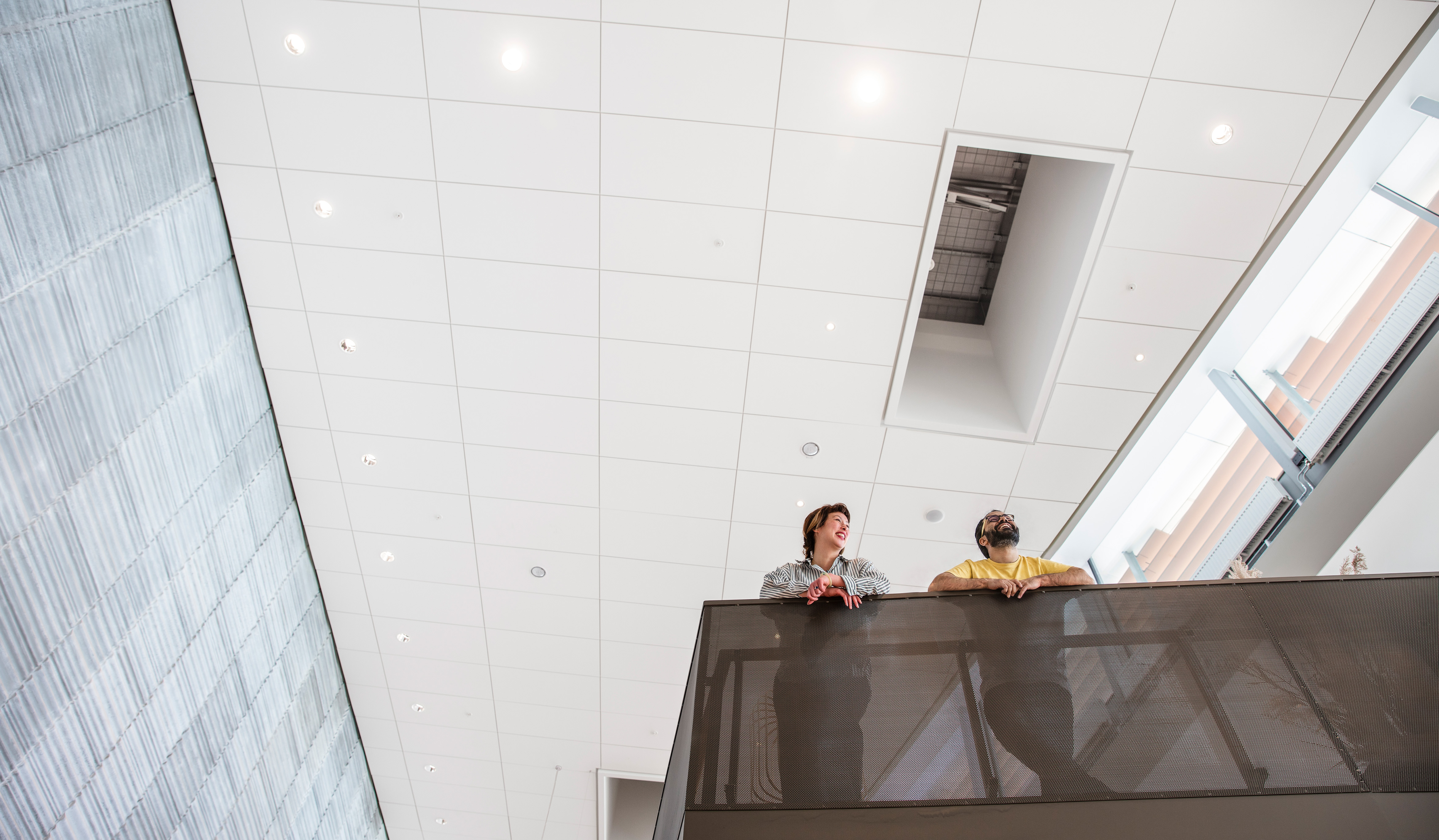 Two people stand and talk on an indoor balcony, viewed from below. The ceiling above them is white with many lights, and the space feels open and modern.