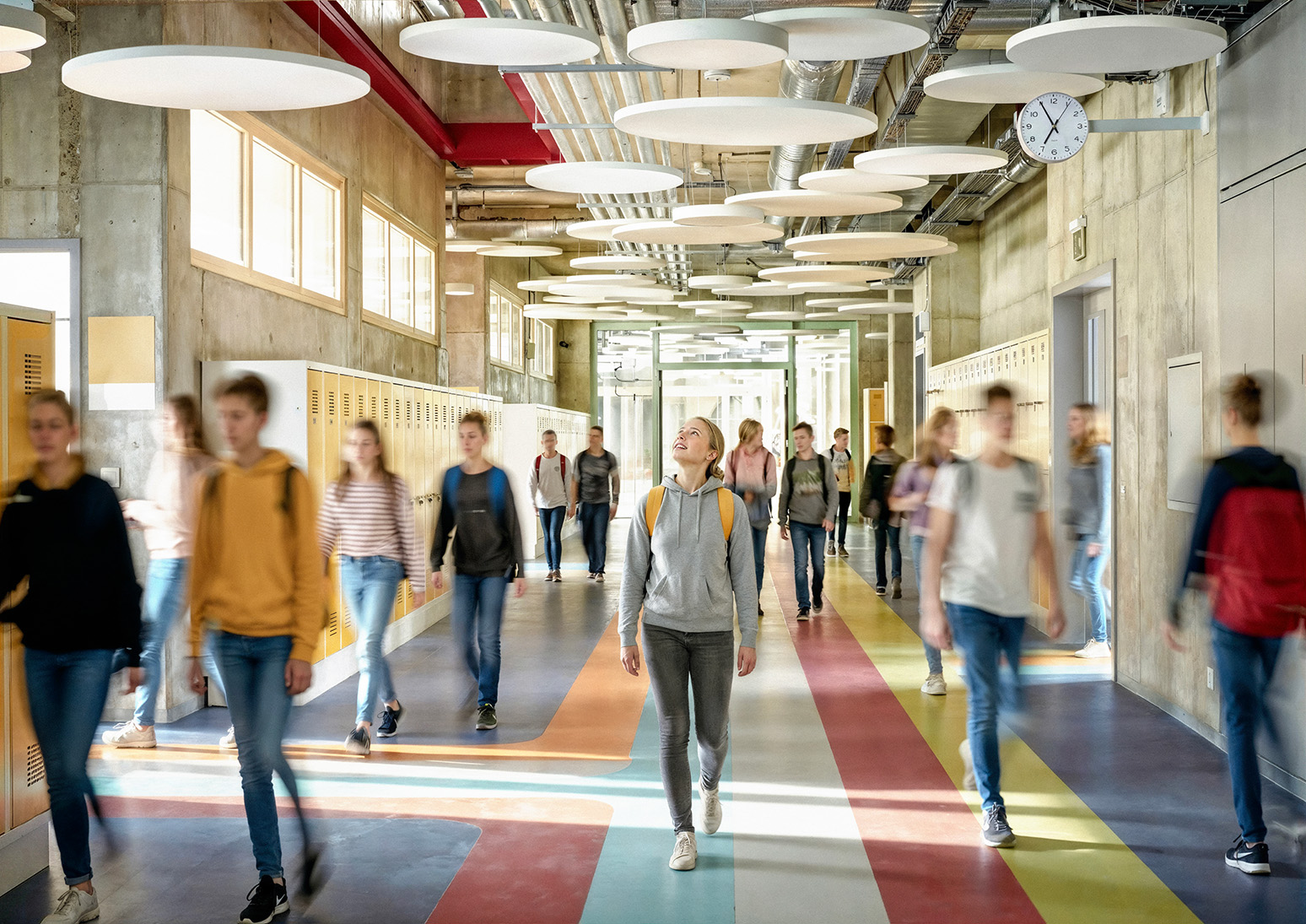 Students walk through a brightly lit school corridor with colourful floors, yellow lockers, and circular ceiling panels; one pupil looks up while others move around. A clock on the wall shows the time.