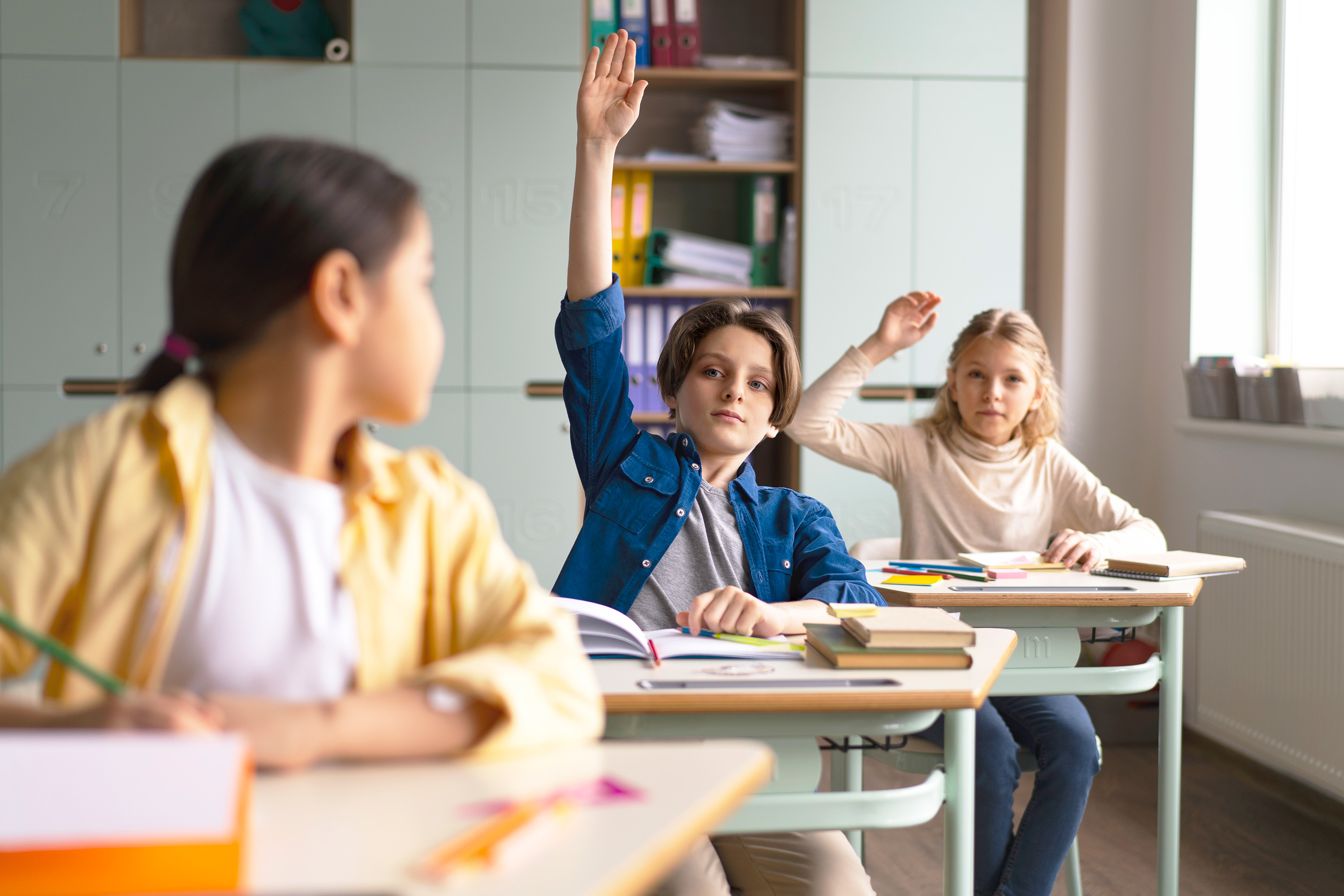 Three children sit at desks in a classroom. Two children in the background have their hands raised, whilst the child in the foreground looks towards them. Books and school supplies are on their desks.