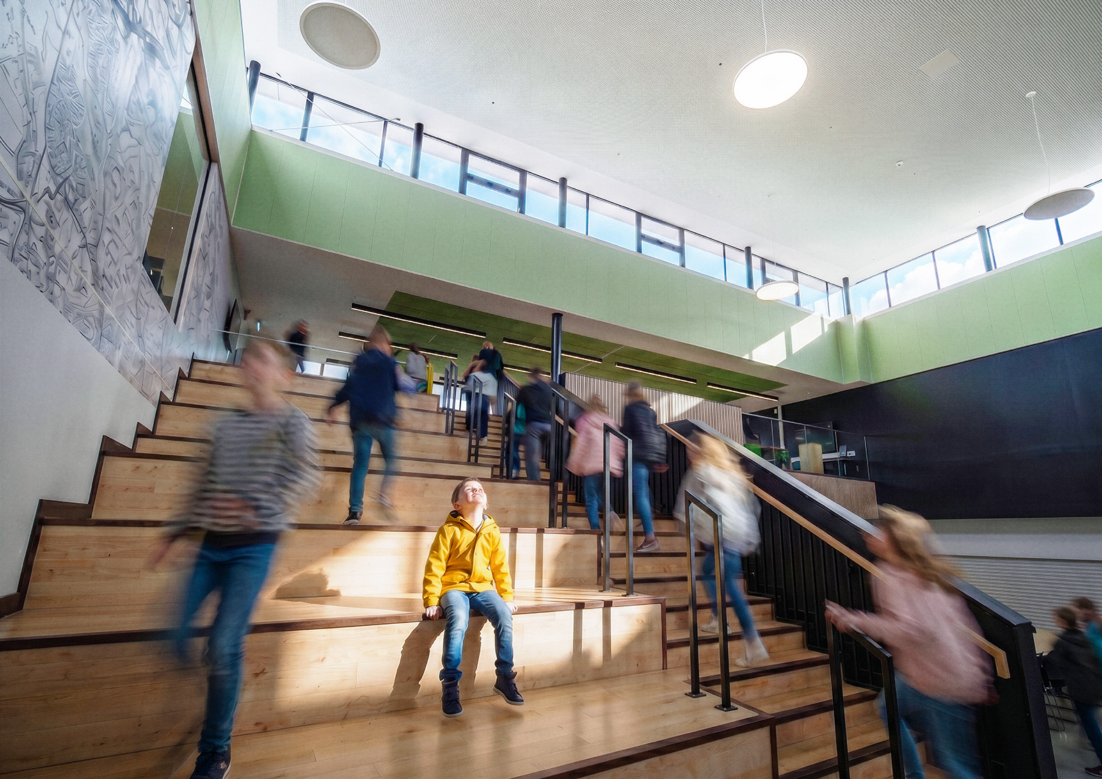 A child in a bright yellow jacket sits on wide wooden steps in a modern, sunlit building, whilst others move quickly up and down the stairs, creating a blur of motion around them.