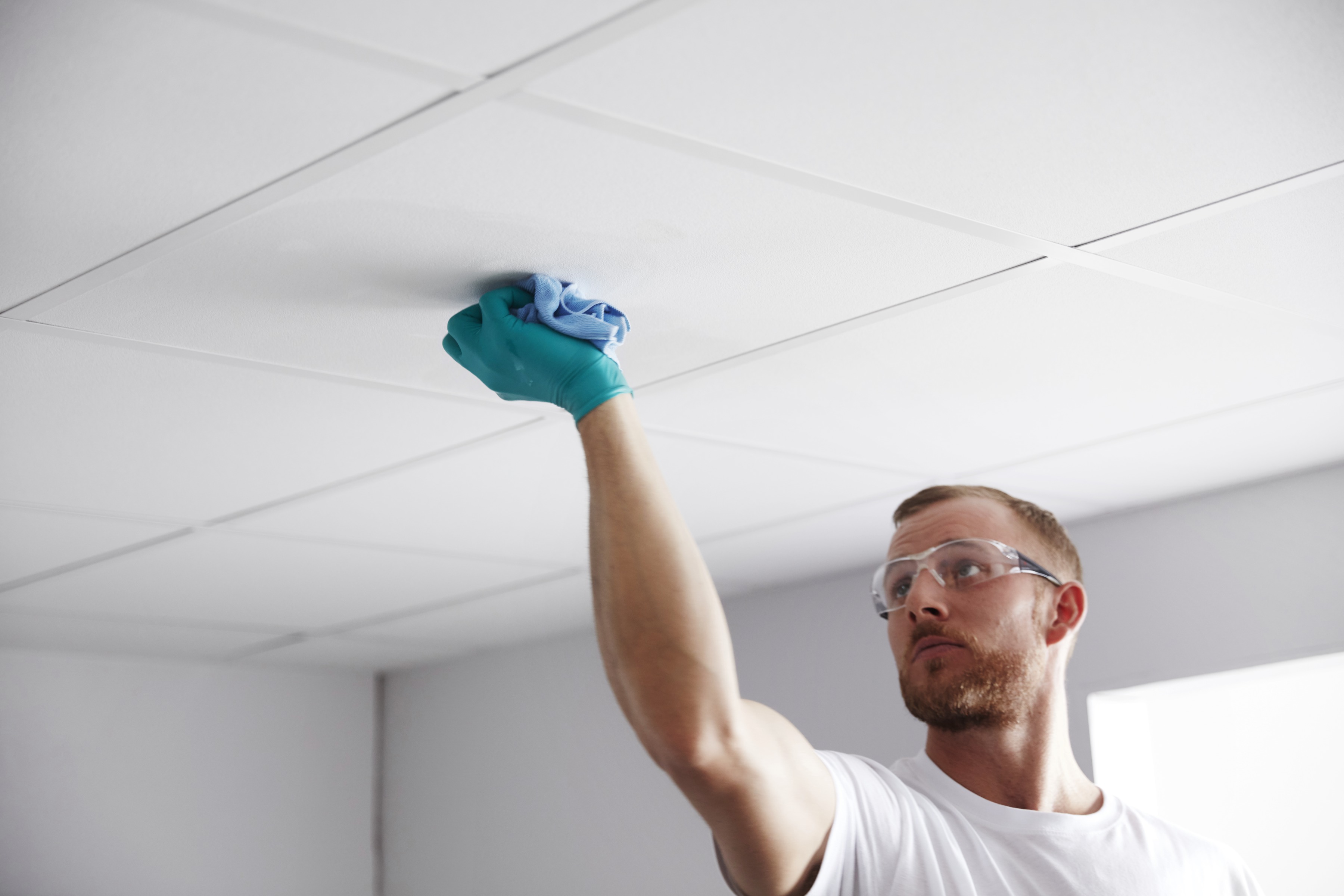 A man wearing safety goggles and a glove cleans a white ceiling tile with a blue cloth in a brightly lit room.