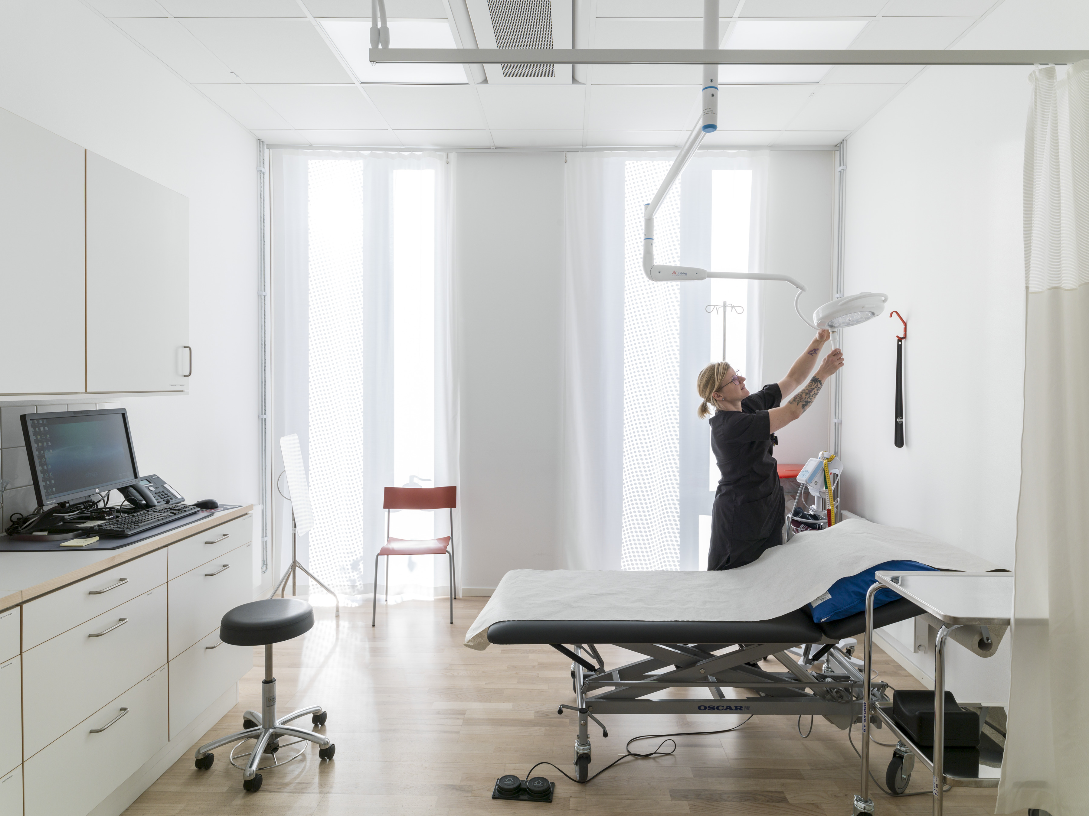 A medical examination room with a bed, stool, and computer. A person adjusts an overhead examination light. The room is clean, bright, and minimal, with white walls, wooden floors, and large windows.