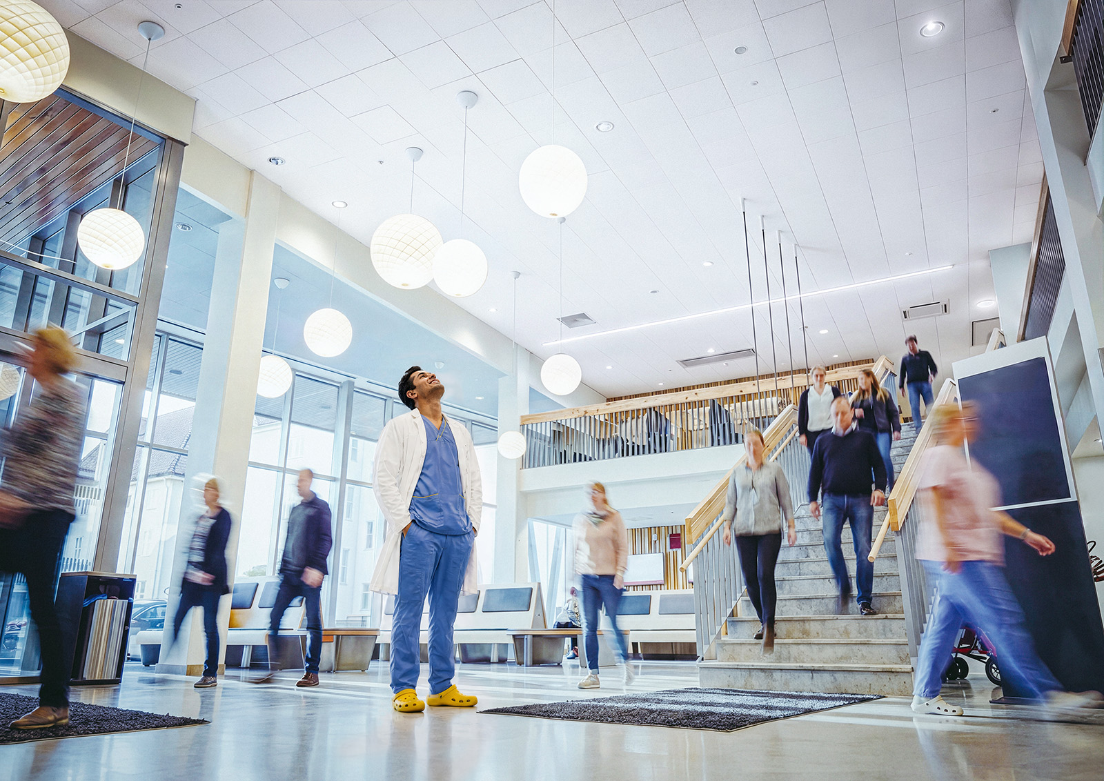 A healthcare worker in scrubs and a white coat stands still in a busy hospital foyer as people walk quickly around him. The foyer is bright with natural light, modern decor, and round ceiling lights.
