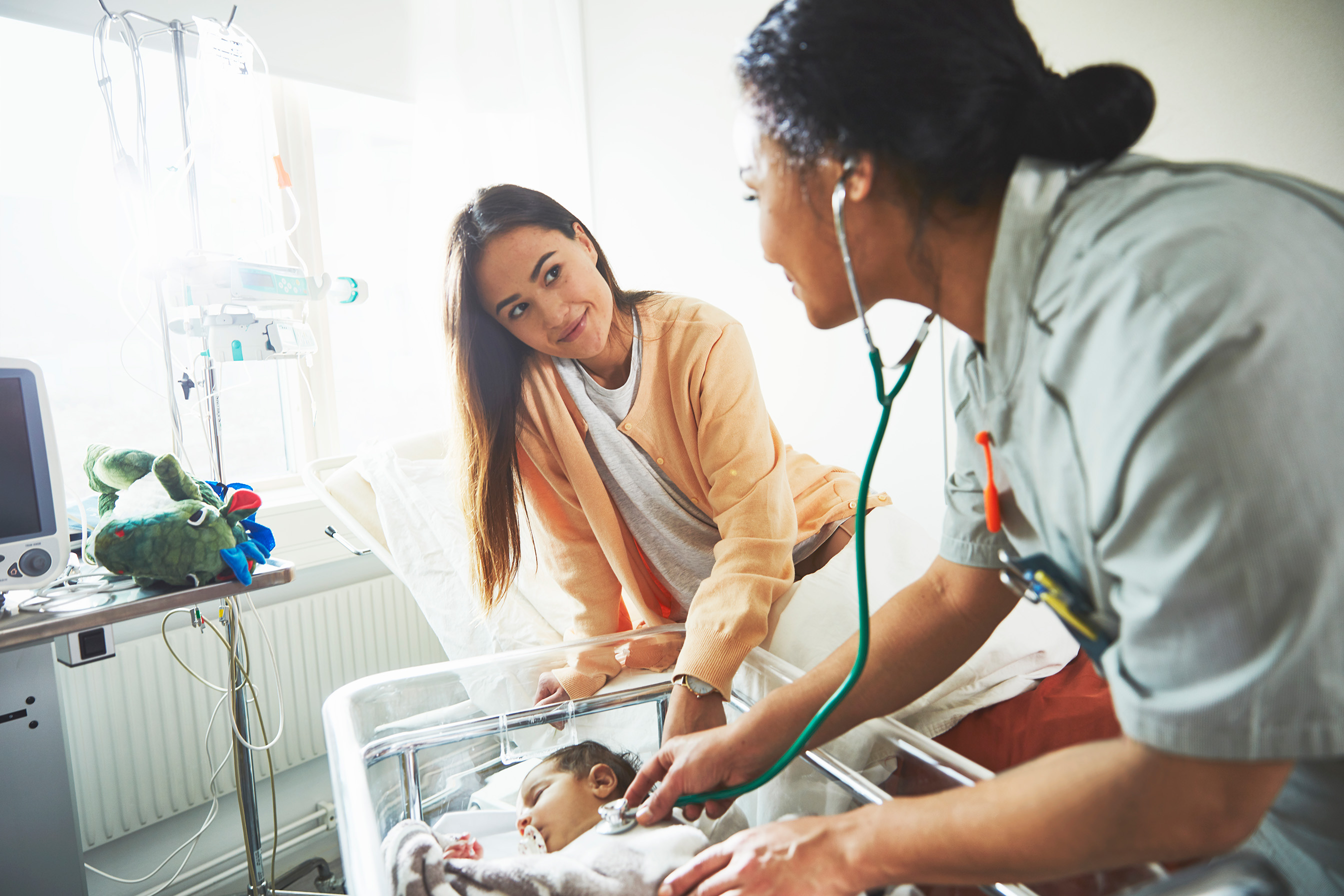 A nurse uses a stethoscope to check a newborn baby lying in a hospital cot, while the baby’s mother watches attentively, resting her hand on the cot. Medical equipment and a soft toy are nearby.