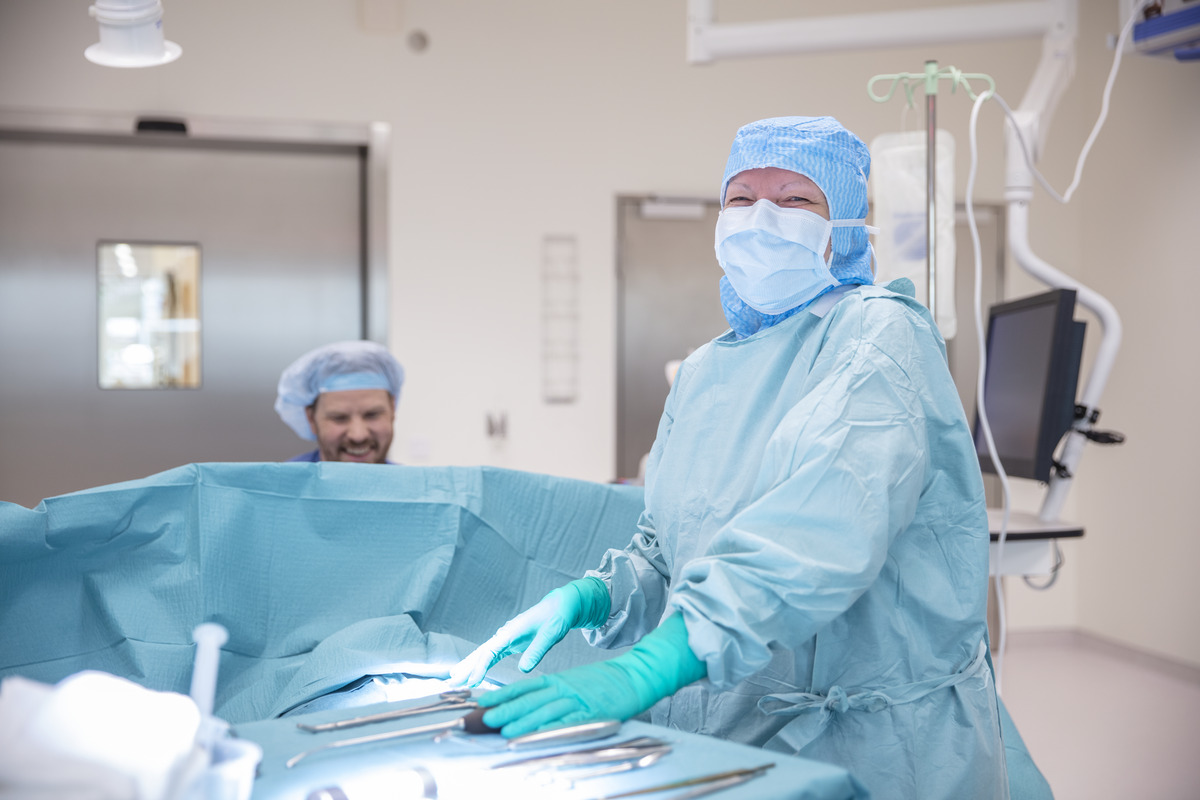 A surgeon in a blue gown, mask, and gloves stands in an operating theatre, smiling while preparing surgical instruments. Another medical professional, also in scrubs, is seated in the background.