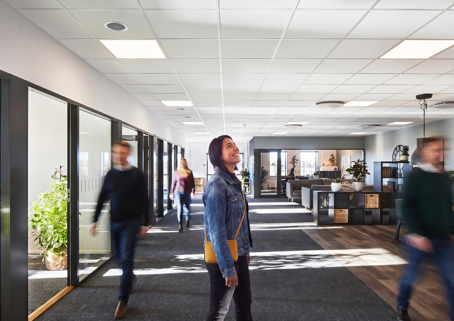 A woman in a denim jacket stands and looks up in a bright, modern office corridor, while several people walk around her. The space features glass office walls, indoor plants, and natural light.