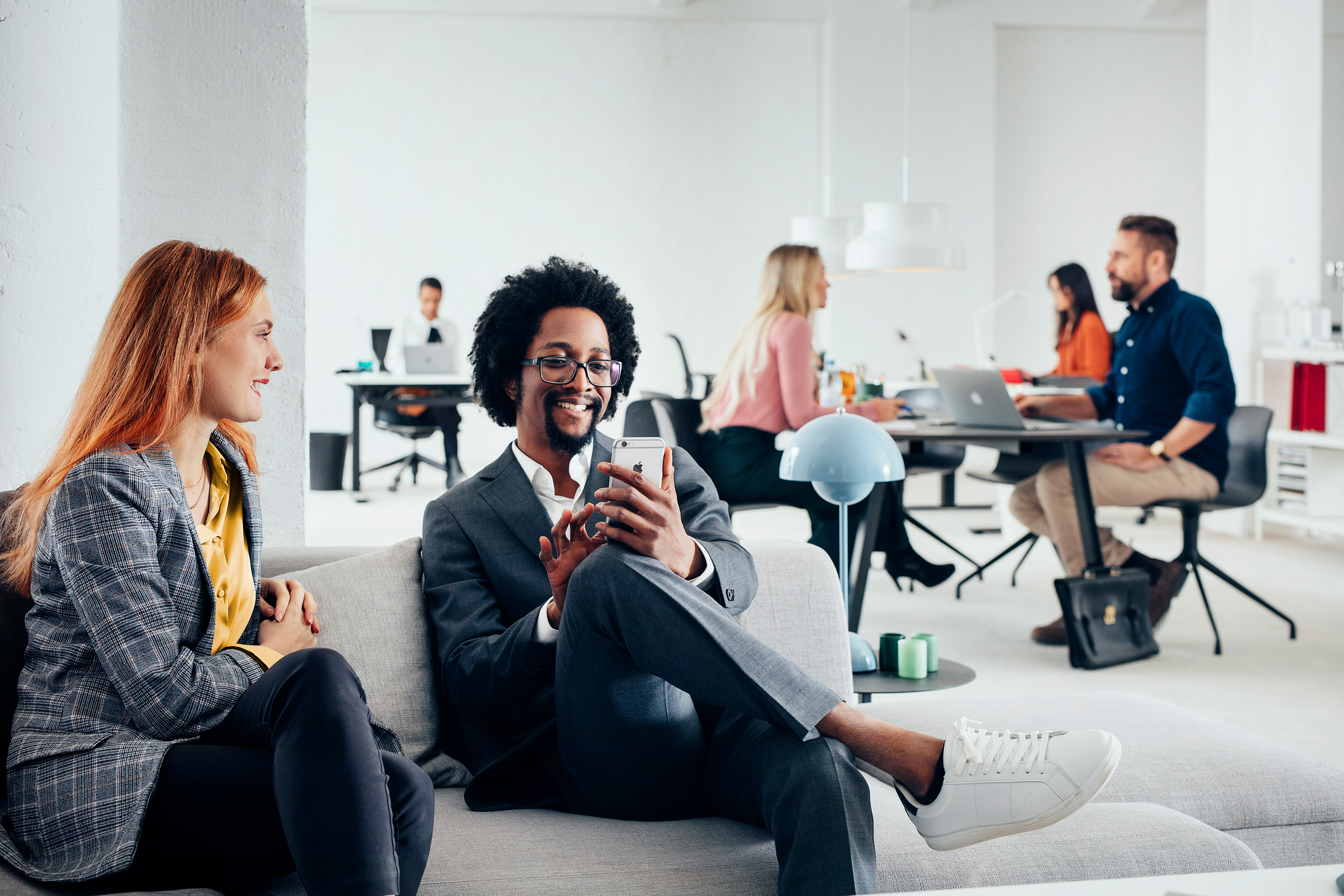 Two colleagues sit on a sofa smiling and looking at a mobile phone, while several people work at desks in a bright, modern open-plan office in the background.