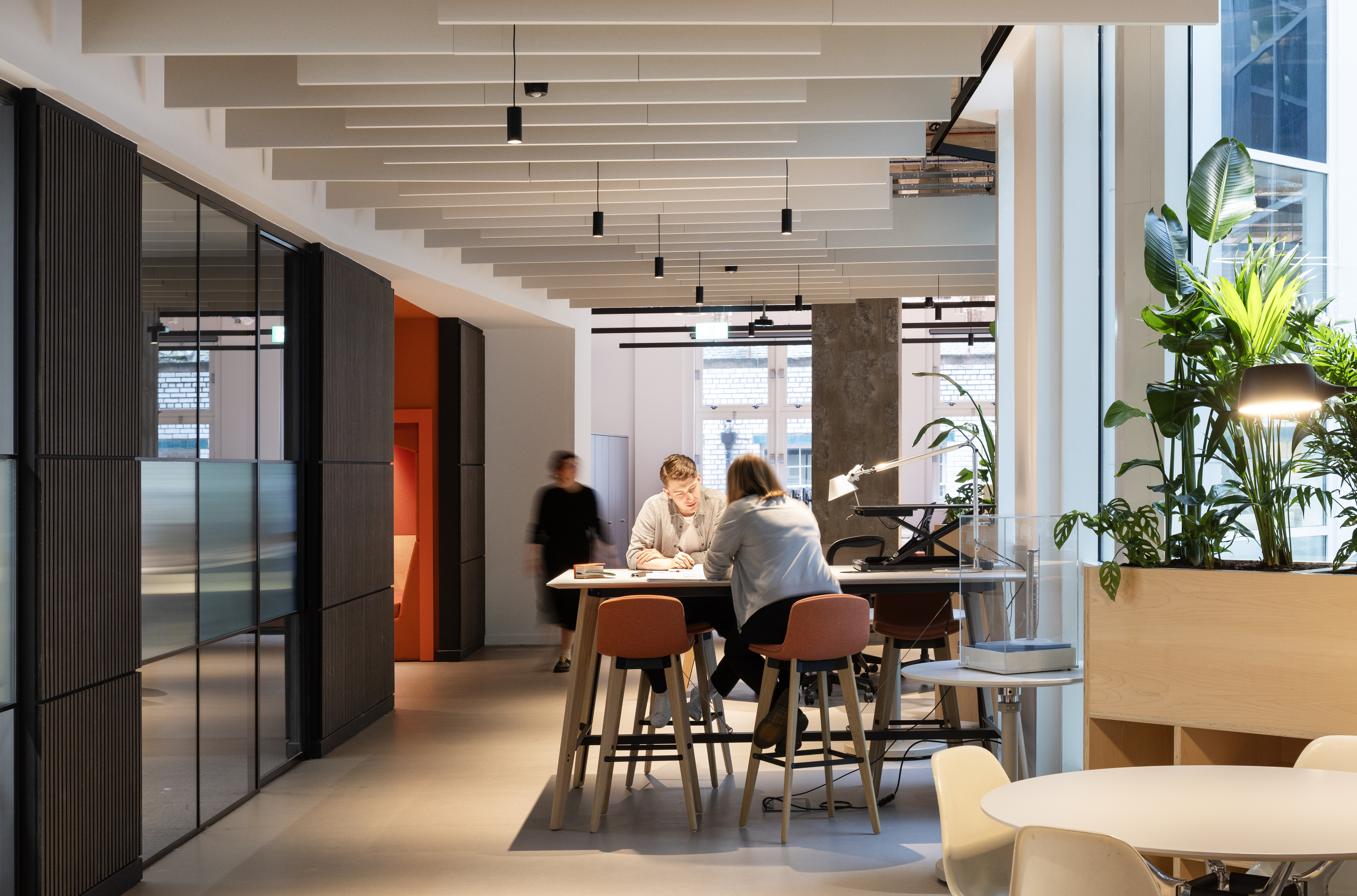 Two people sit and talk at a high table in a modern, brightly lit office space with plants, large windows, and sleek furniture. Another person walks by in the background.