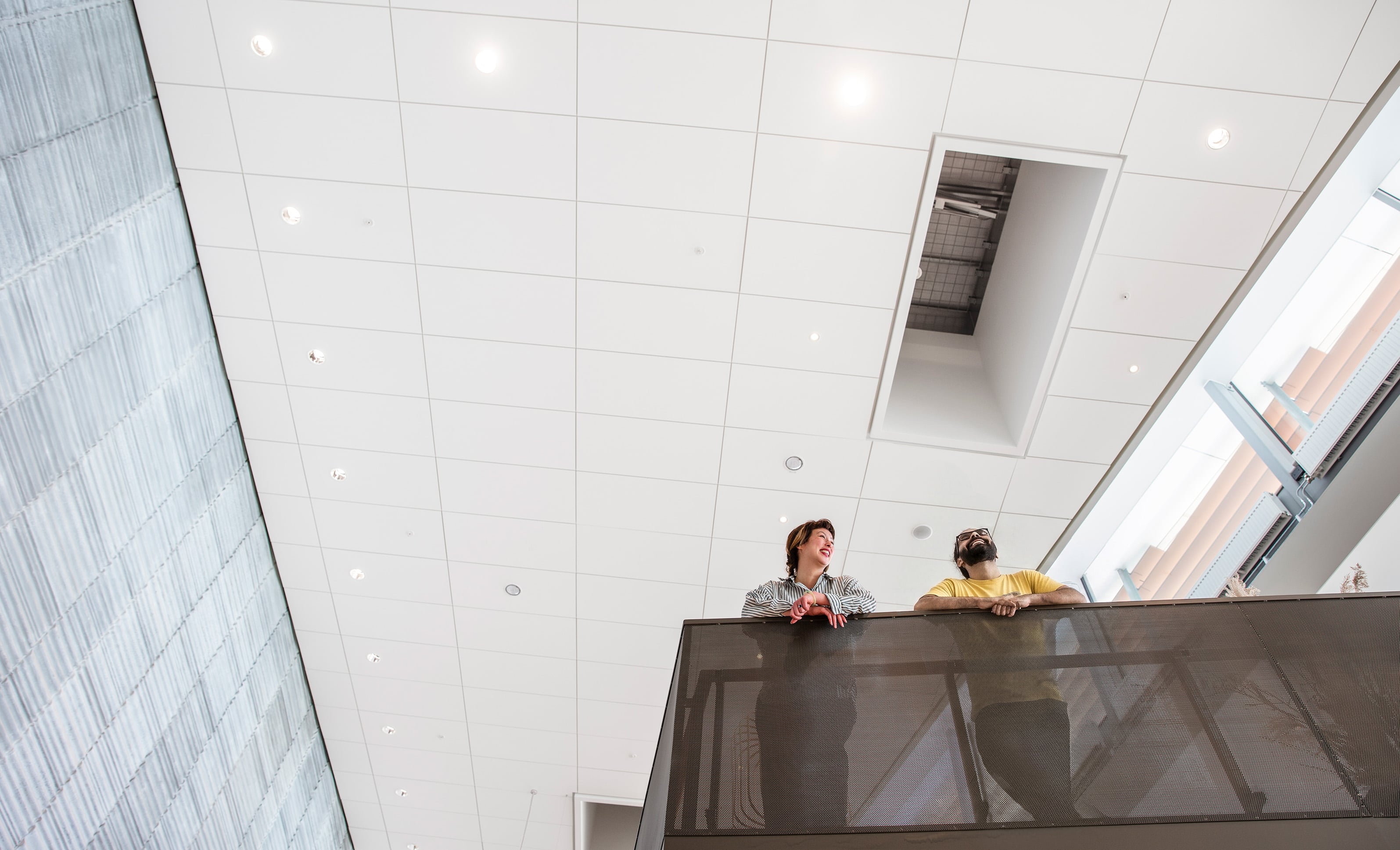 Two people stand and talk on an indoor balcony, viewed from below. The ceiling above them is white with many lights, and the space feels open and modern.