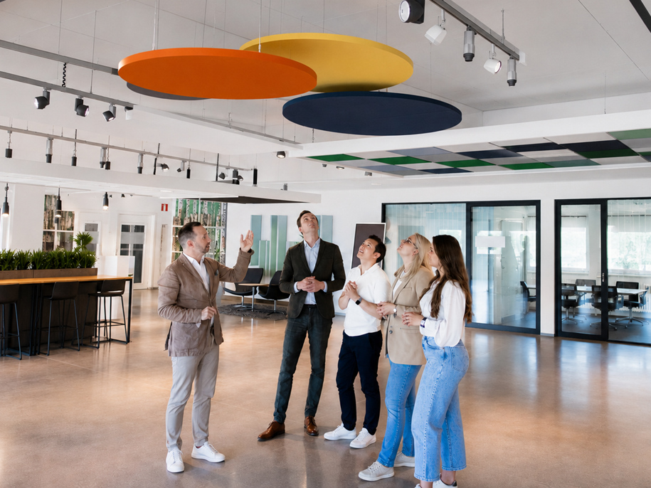 Five people stand in a modern office space, looking up at large colourful circular panels on the ceiling. One person gestures upwards while the others watch. The office has glass walls, plants, and bright lighting.