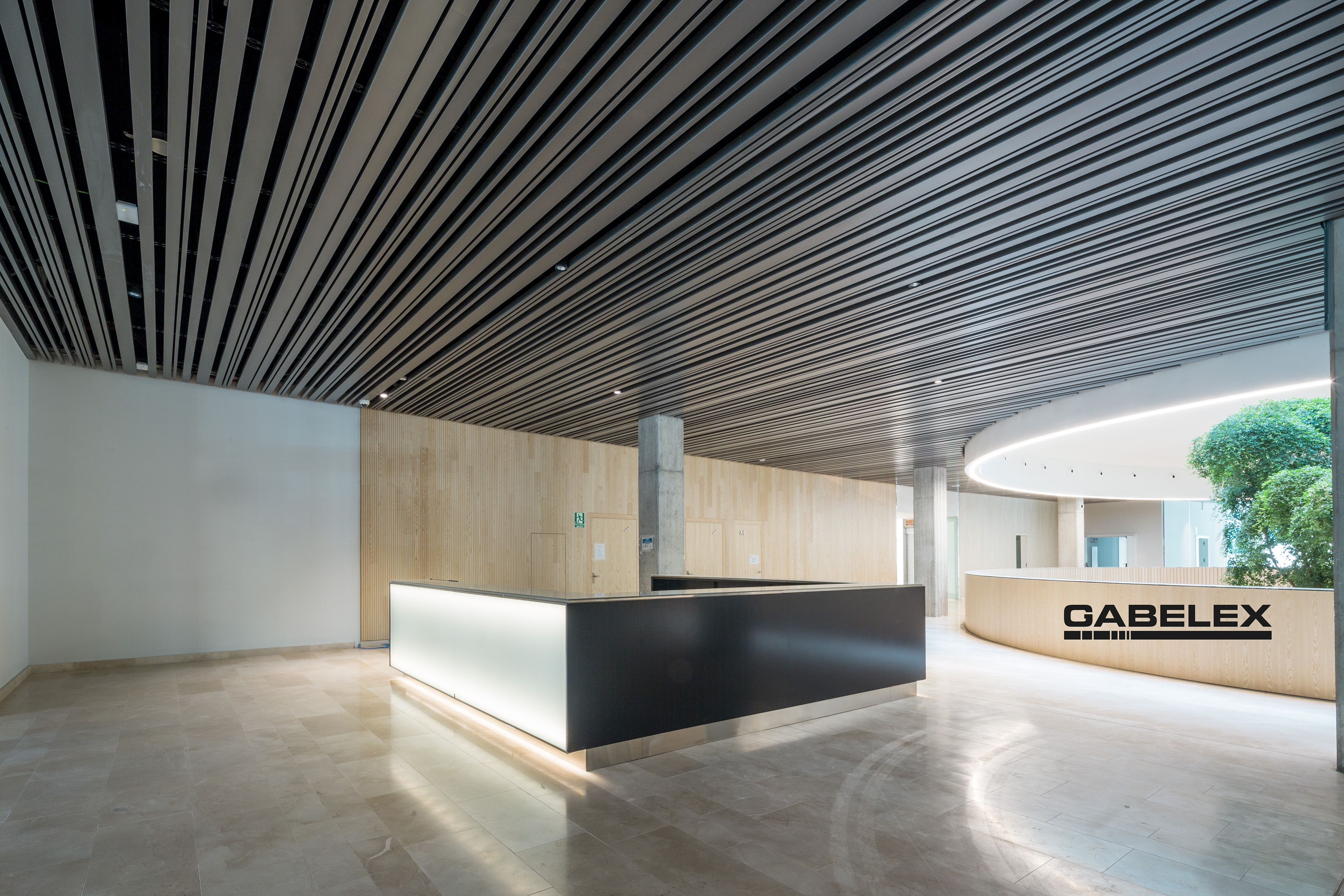 Modern office lobby with a sleek, illuminated reception desk in the center. The ceiling has black slats, and there is a curved wooden wall with the text GABELEX on it. A round space with greenery is visible in the background.