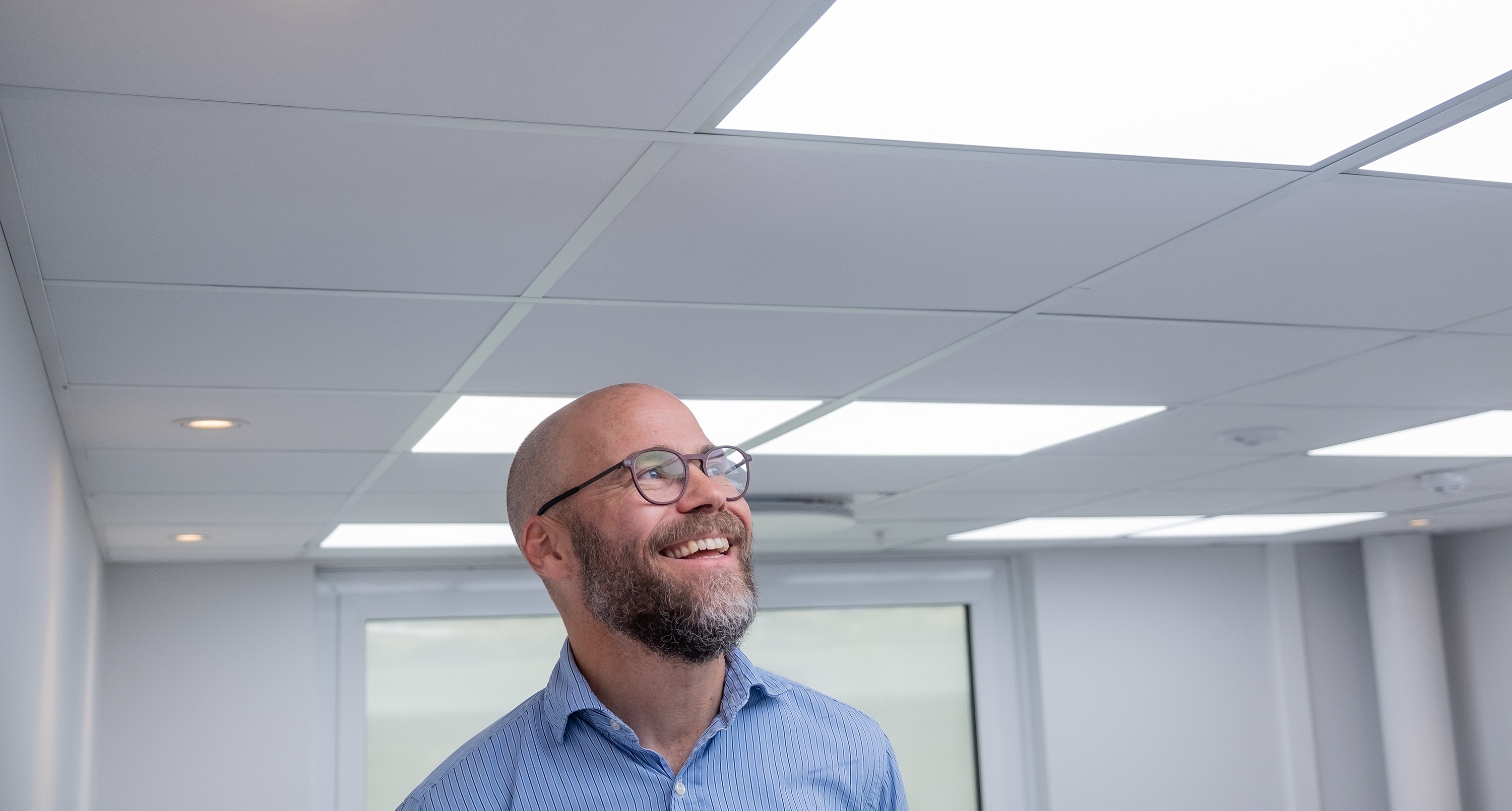 A smiling man with glasses and a beard, wearing a blue shirt, standing in a well-lit room with biocentric ceiling lights.