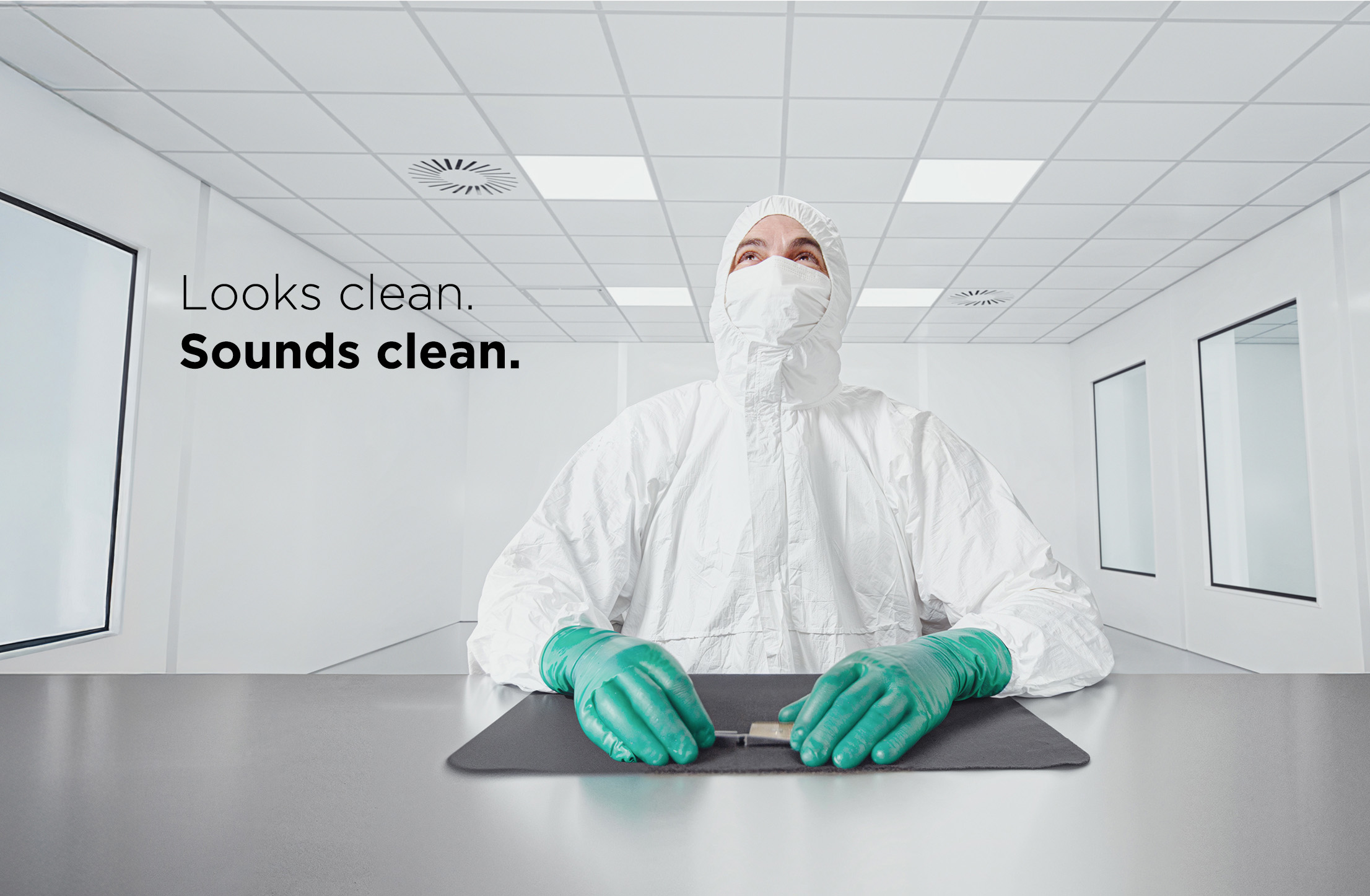 A person in a white hazmat suit and green gloves sits at a table in a clean, sterile room. Text on the wall reads, Looks clean. Sounds clean. The environment appears highly sanitised.
