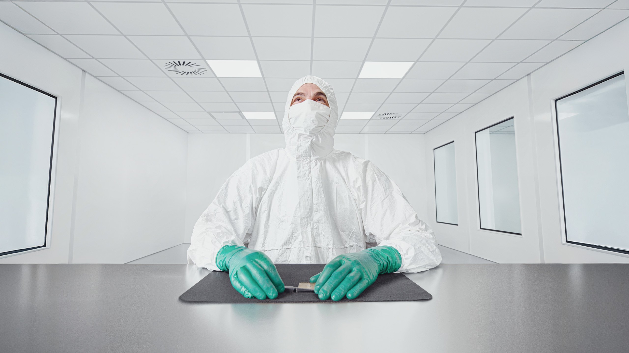 A person in a white cleanroom suit, mask, and green gloves sits at a metallic table in a bright, sterile laboratory, holding a small object on a black mat. Large windows line the white walls.