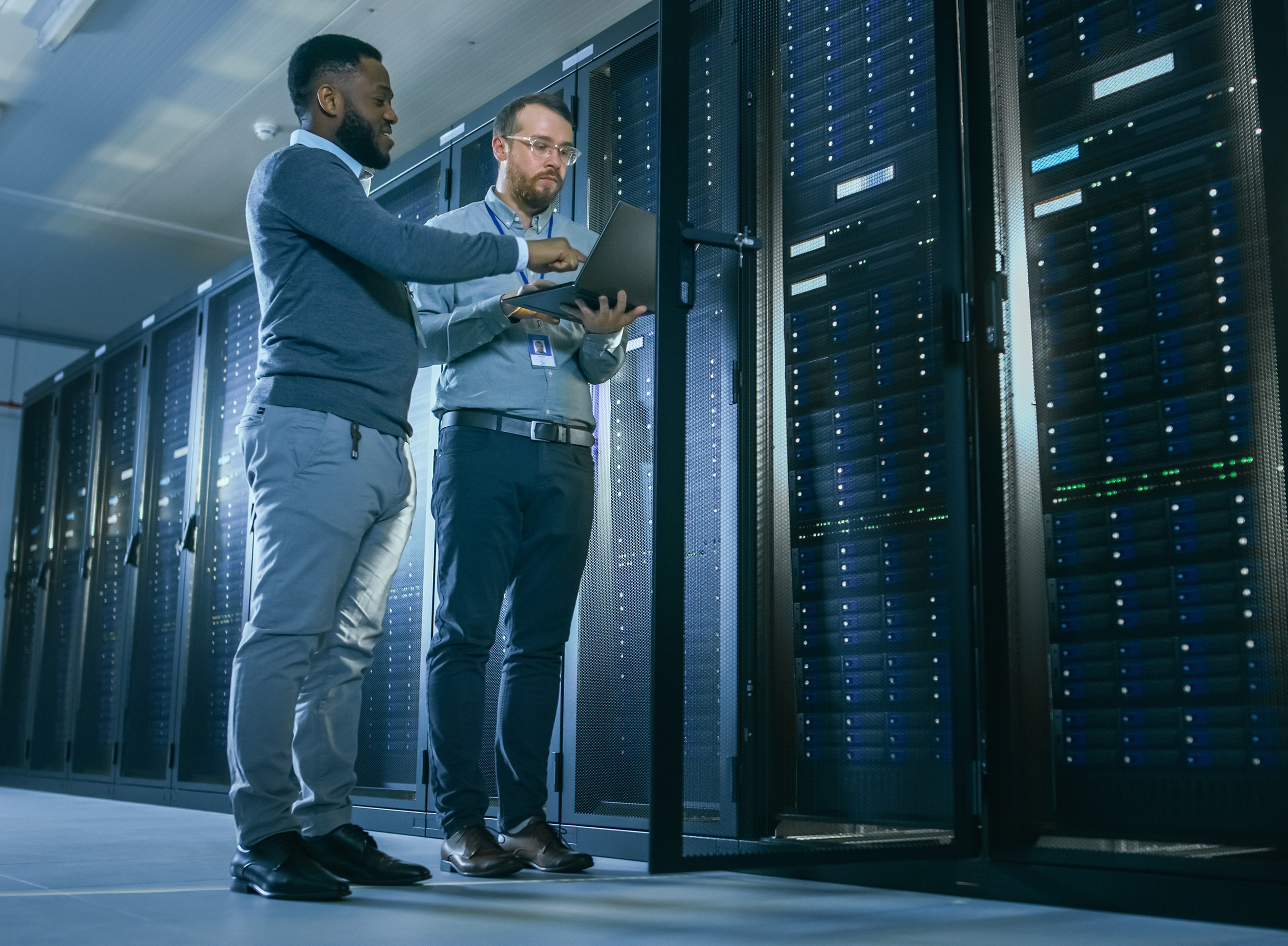 Two men stand in a server room, examining data on a laptop. Tall server racks filled with equipment line the room, and one rack door is open. The environment appears clean, modern, and professional.