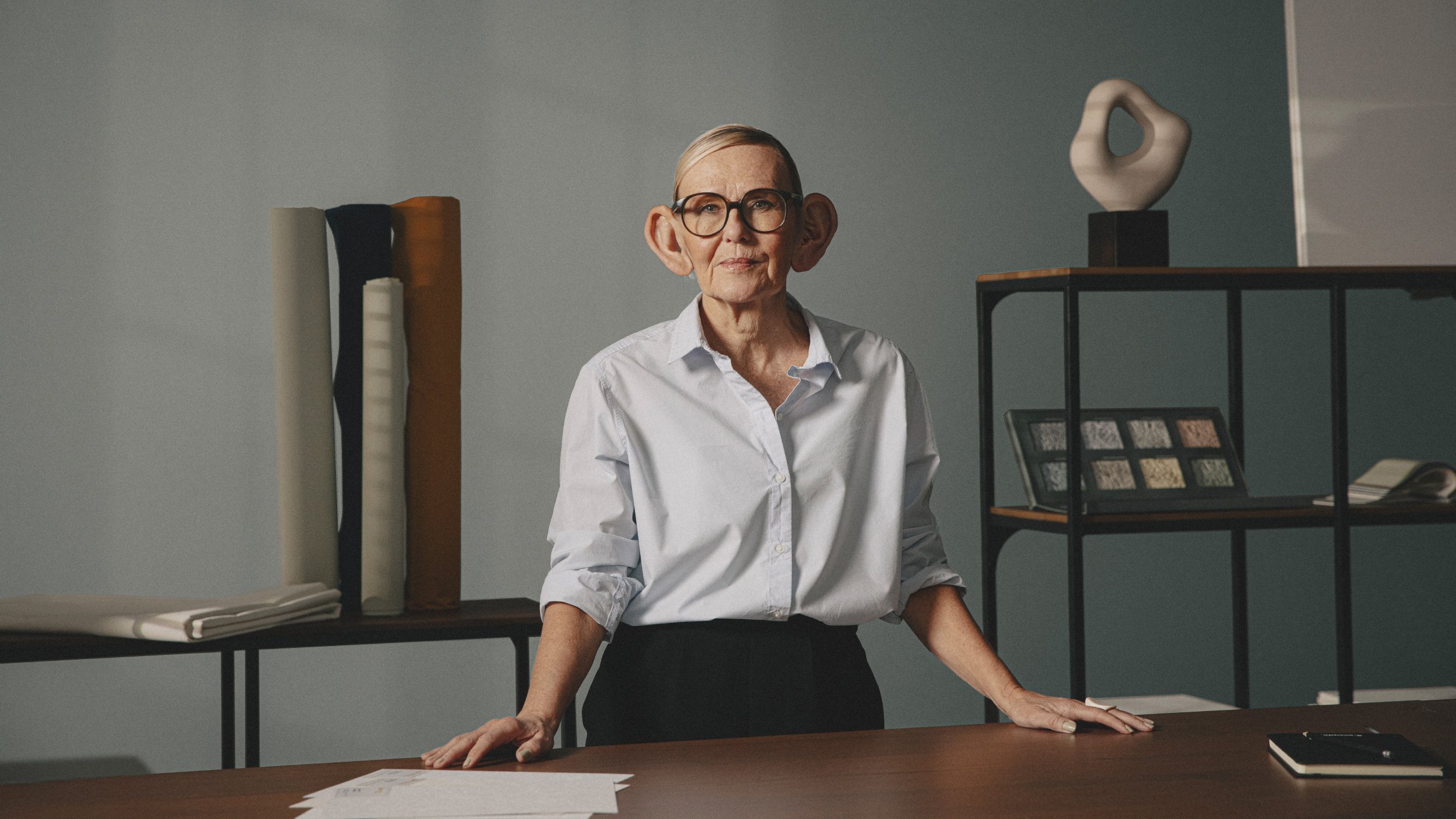 A person with oversized, cartoonish ears stands confidently at a table with acoustic design materials. The background shows shelves with various design objects.