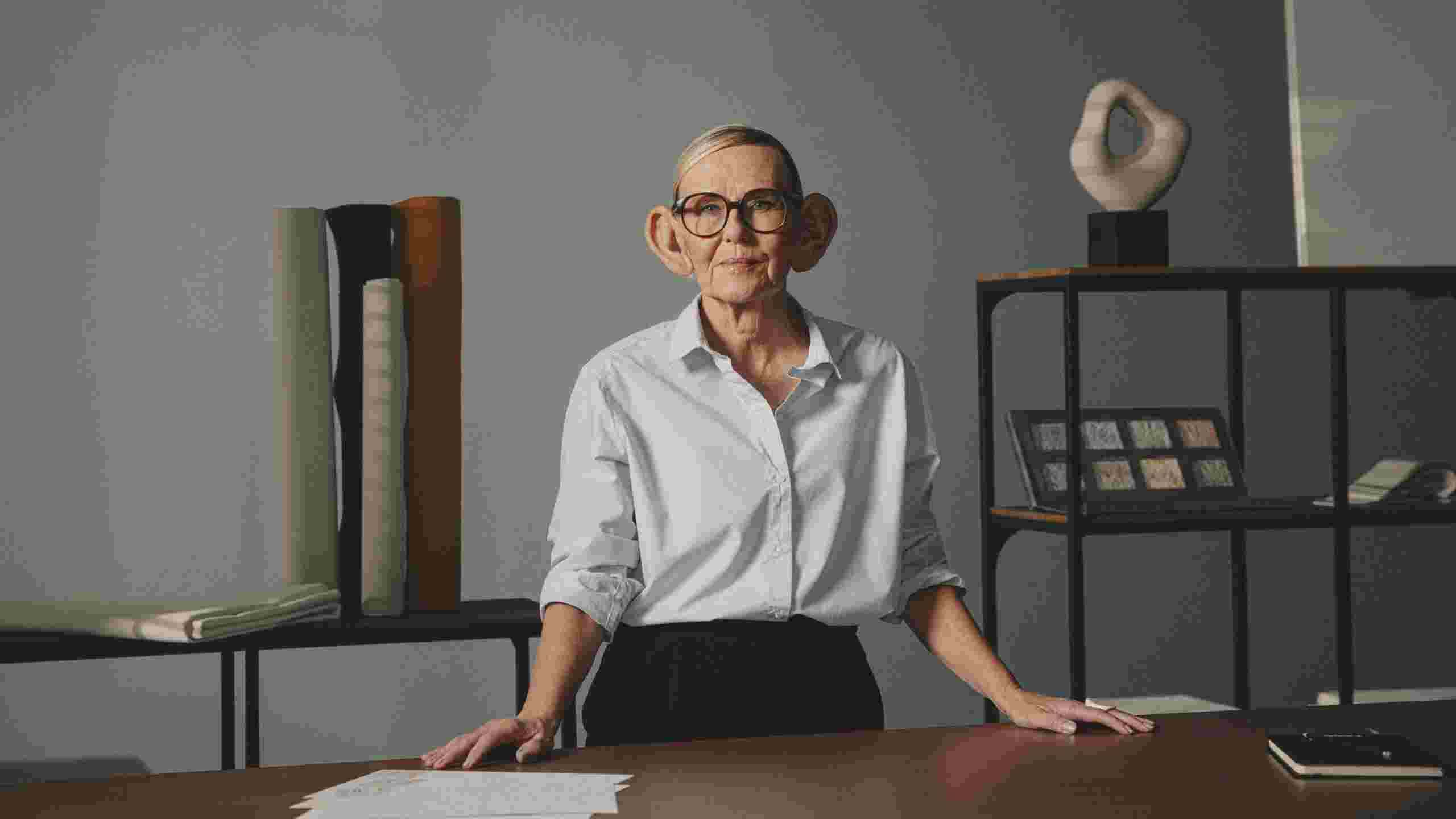 A person with oversized, cartoonish ears stands confidently at a table with acoustic design materials. The background shows shelves with various design objects.