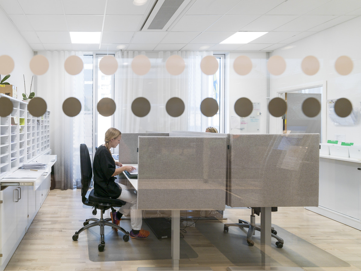 Two people work at desks in a bright office with large windows and frosted glass dots. One person types on a computer, shelves and office supplies are visible in the background.