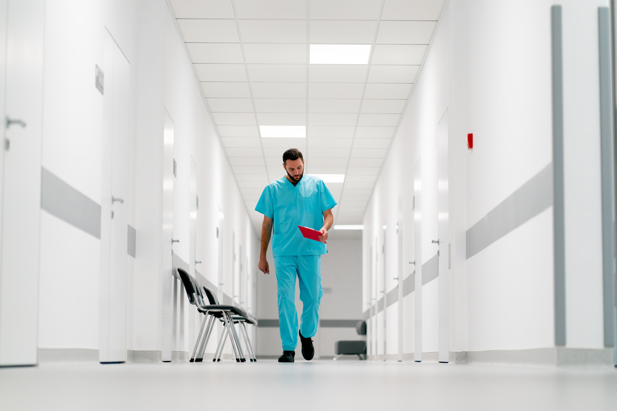 A healthcare worker in blue scrubs walks down a bright, empty hospital corridor, looking at a red folder. Several black chairs are lined up against the wall on the left.