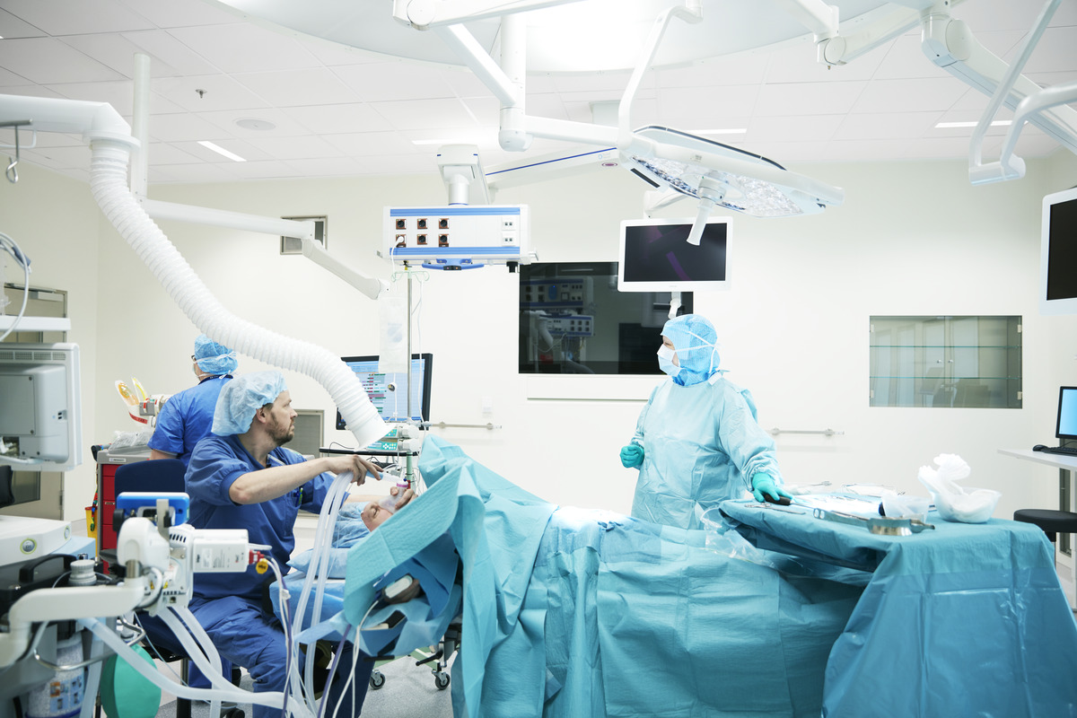 Surgeons and medical staff in blue scrubs perform an operation in a brightly lit, modern operating theatre filled with medical equipment and monitors.