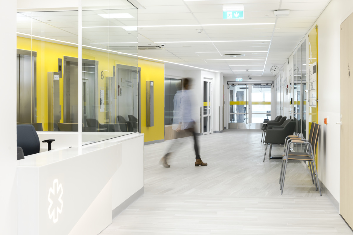 Modern hospital corridor with yellow accents, empty seating, glass walls, and a blurred person walking past a reception desk. Bright lighting and clean, minimalist décor.