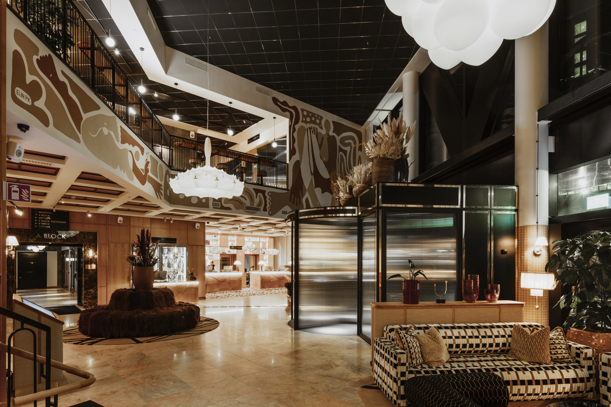 Modern hotel lobby with warm ambient lighting. The space features a mix of contemporary and art deco design elements, including a patterned acoustic ceiling, decorative wall panels with abstract designs, and stylish furniture. A central seating area with plush sofas and chairs is arranged around a circular wooden table adorned with large feathered plants. To the right, there is an elevator with reflective doors next to potted plants. The lobby extends into the background where more seating areas and hotel amenities are visible under soft, glowing light fixtures that resemble clouds. 