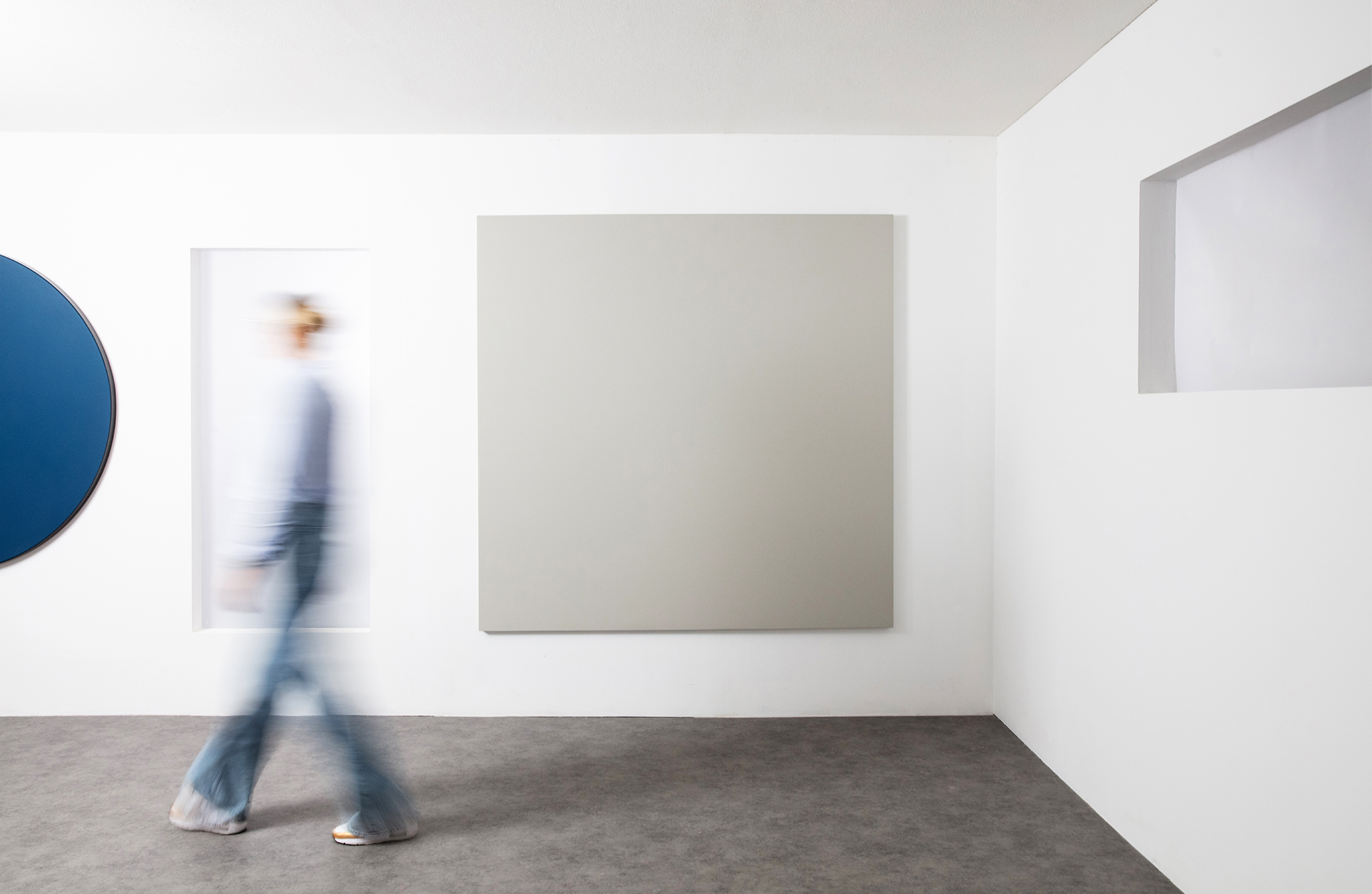 A person in motion wearing jeans and a sweater walks past a minimalist wall. The wall features a large grey square and a circular blue panel. The setting is modern with white walls and a grey floor.