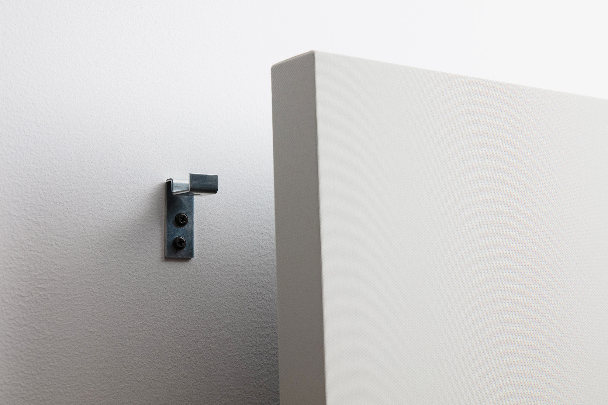 Close-up of a metal wall hook installed on a textured white wall, adjacent to the edge of a large, unframed rectangular canvas with a similar white texture.