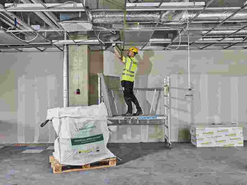 A man standing on scaffolding in an unfinished room demounting acoustic panels for recycling.