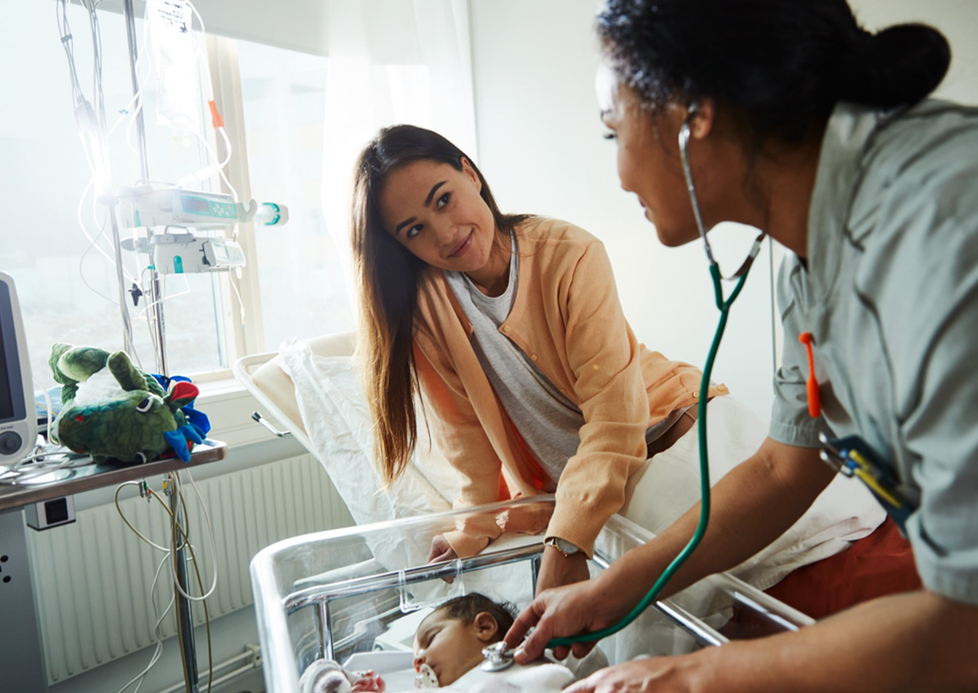 Eine Pflegekraft benutzt ein Stethoskop, um ein Neugeborenes zu untersuchen, das in einem Krankenhausbett liegt, während eine erwachsene Frau, vermutlich die Mutter, lächelnd am Bett des Babys zusieht.