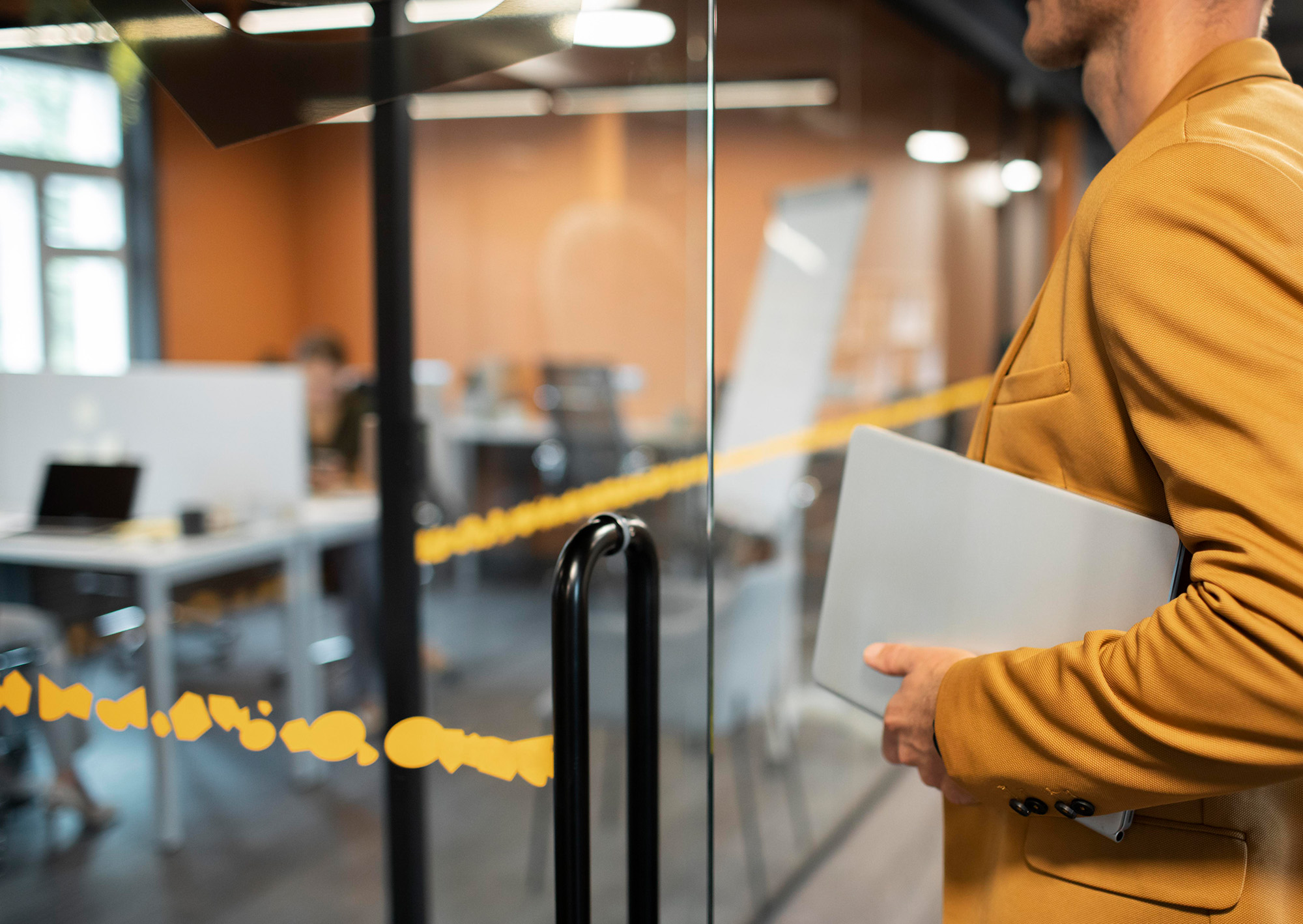A person in a mustard-coloured suit holds a closed laptop while entering a modern glass-walled office with desks and chairs visible in the background.