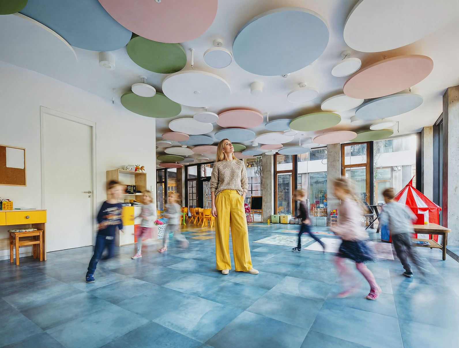 A teacher stands in a colourful, modern classroom with round ceiling panels as young children run and play around her, creating a lively, blurred motion effect.