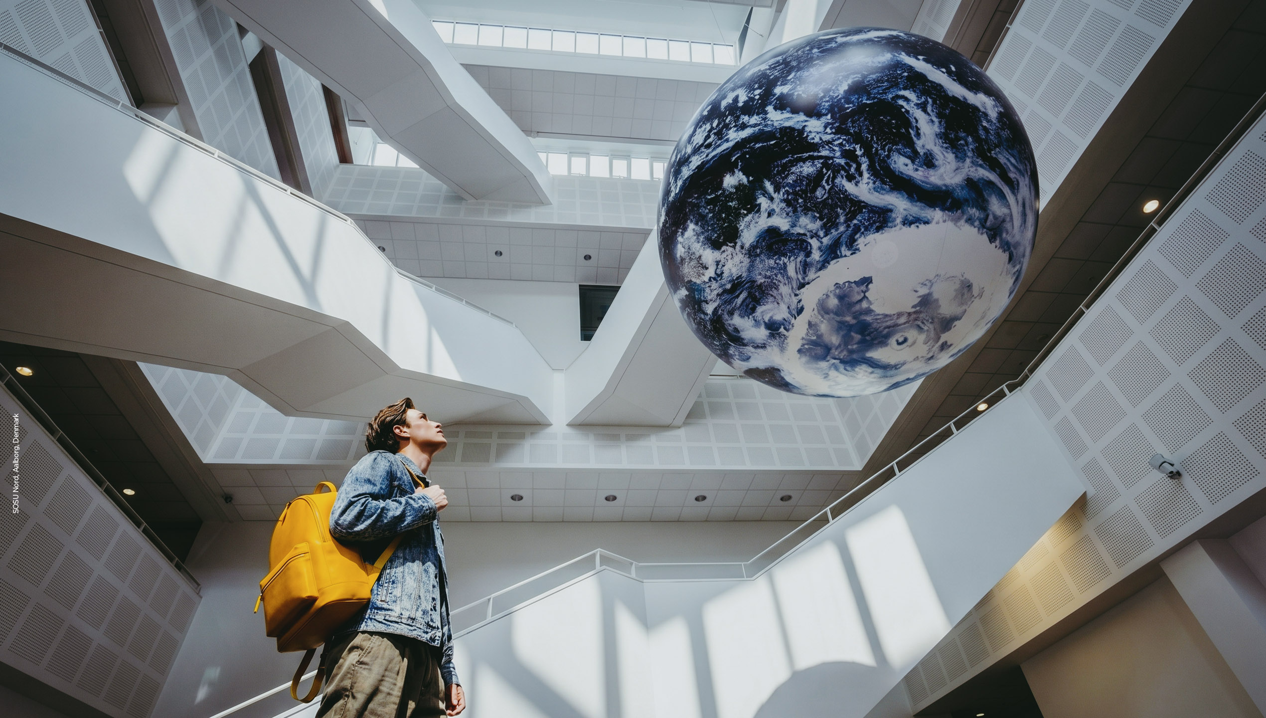 A person with a yellow rucksack looks up at a large globe of Earth suspended in a modern, white architectural space with geometric lines.