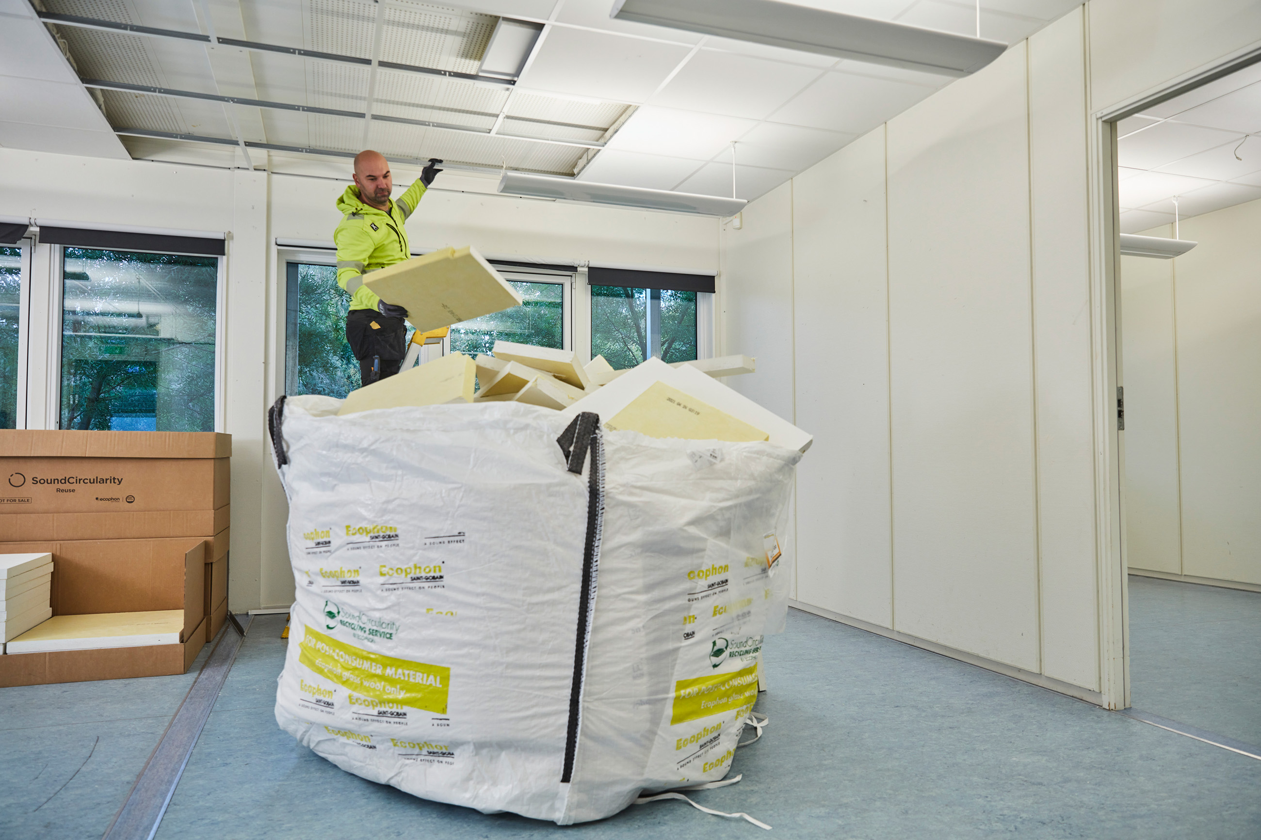 Person removing used acoustic ceiling tiles and placing them in a large recycling bag inside a room during ceiling refurbishment.