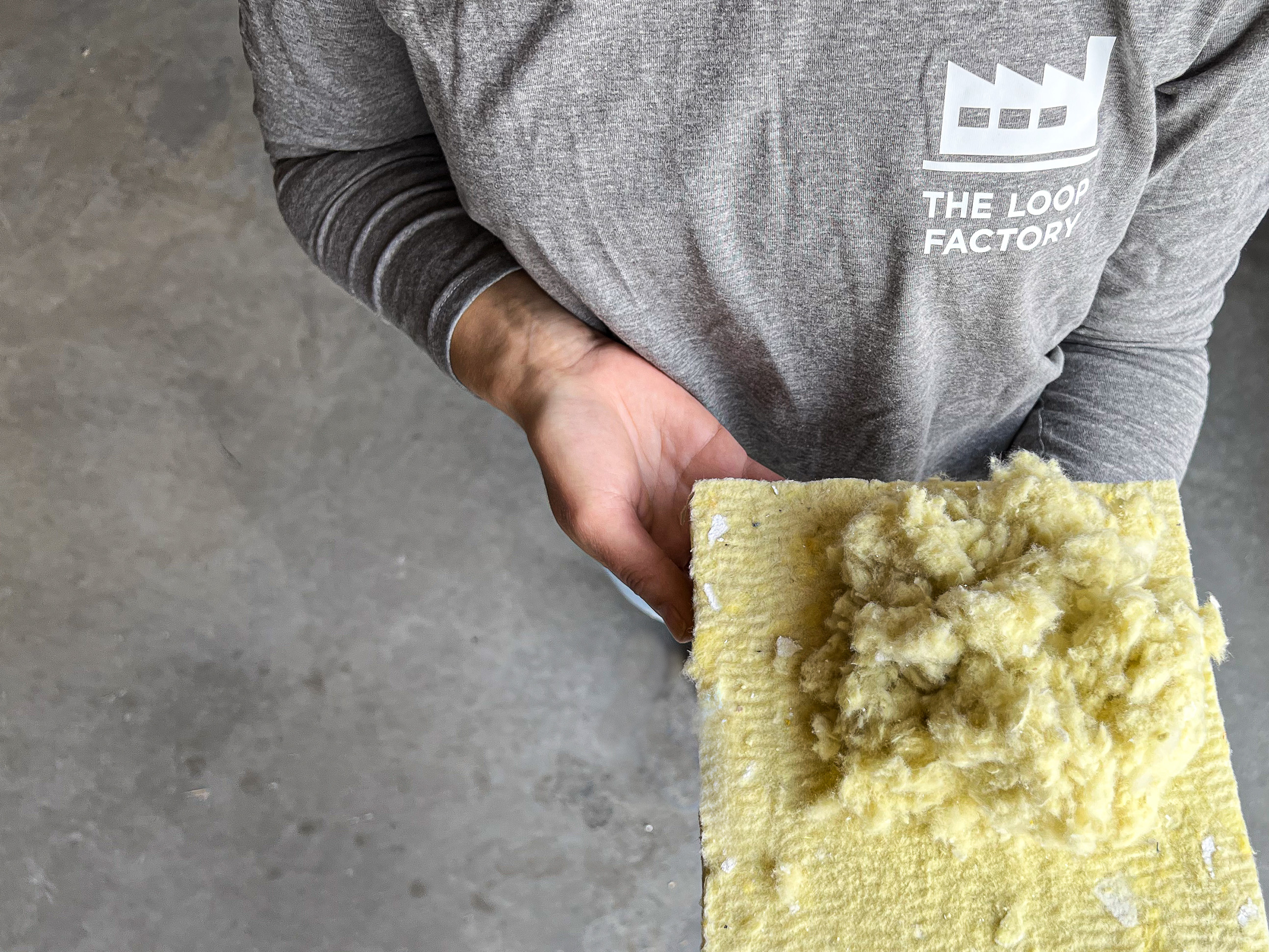 Person wearing a grey shirt with The Loop Factory logo holds a yellow fibrous insulation material in their hand, standing on a grey concrete floor.