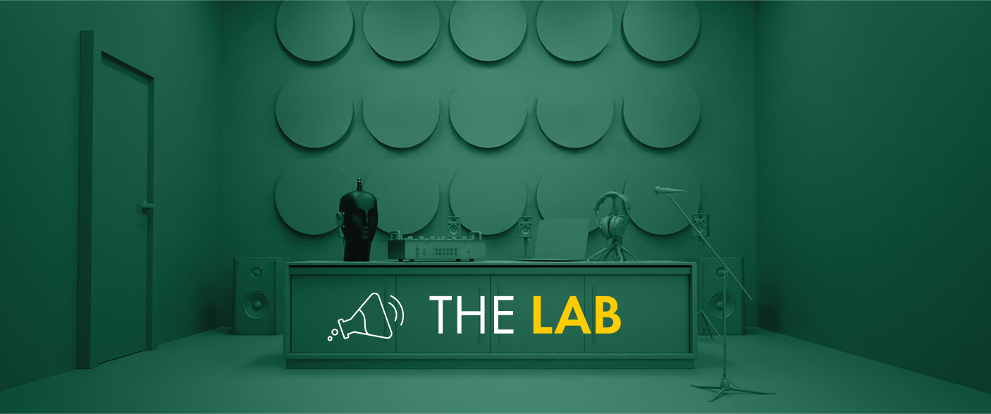 A modern, monochromatic green room with a desk labelled THE LAB in white and yellow letters, featuring sound equipment, a mannequin, speakers, and a microphone on a stand. Circular panels decorate the back wall.